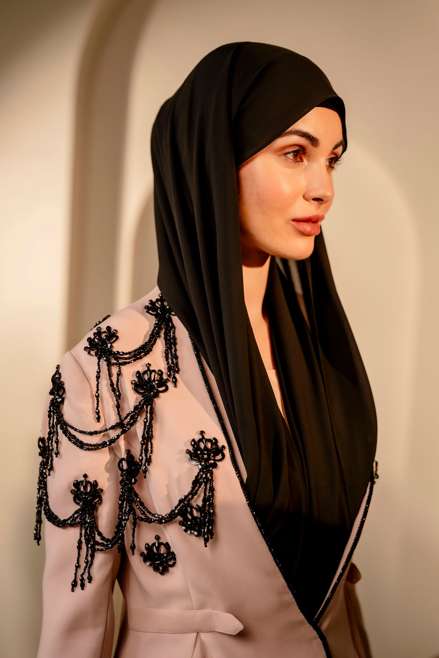 Woman wearing a black hijab and an embellished outfit against a neutral background