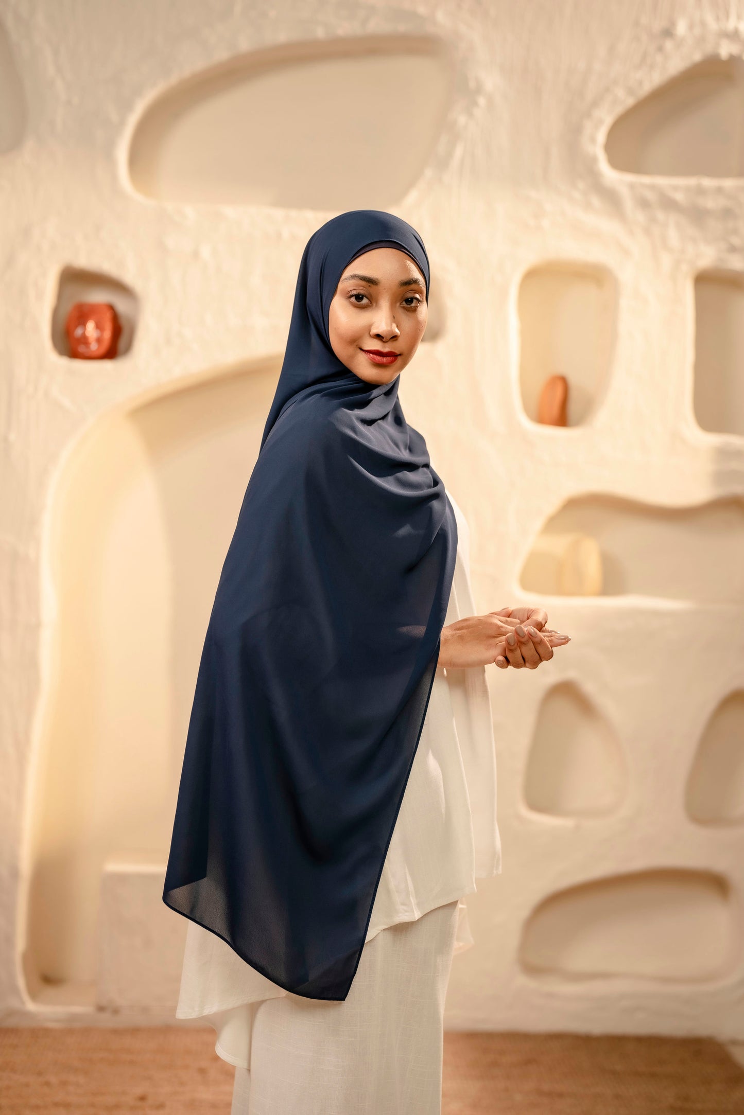 Woman wearing a navy hijab and white dress standing in front of an abstract, light-colored wall
