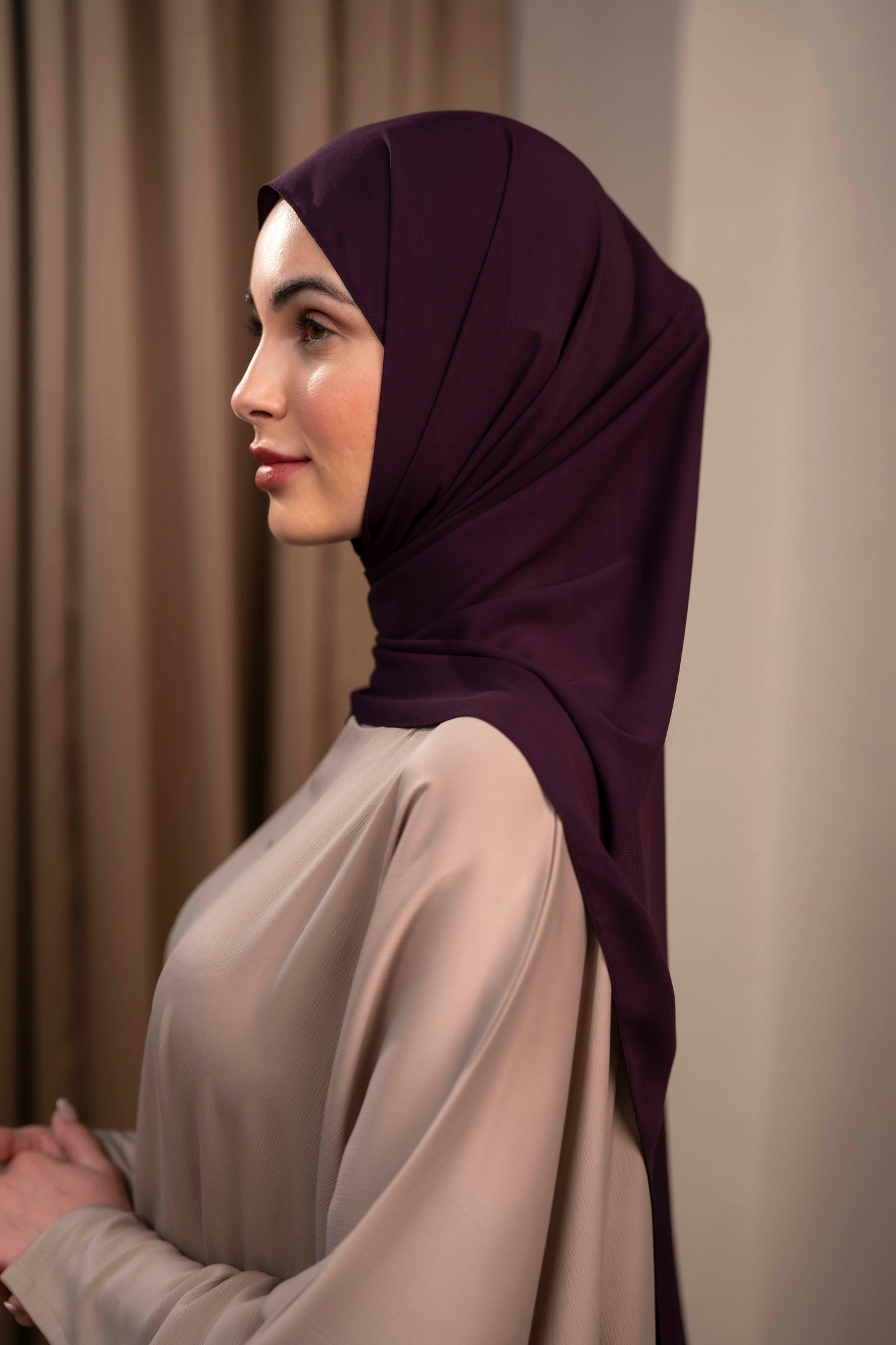 Woman wearing a dark purple hijab and beige top against a neutral background Plum