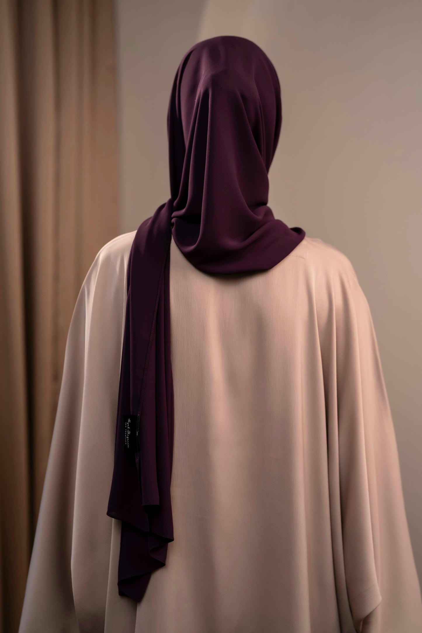 Person wearing a dark purple hijab and beige garment against a neutral background Plum