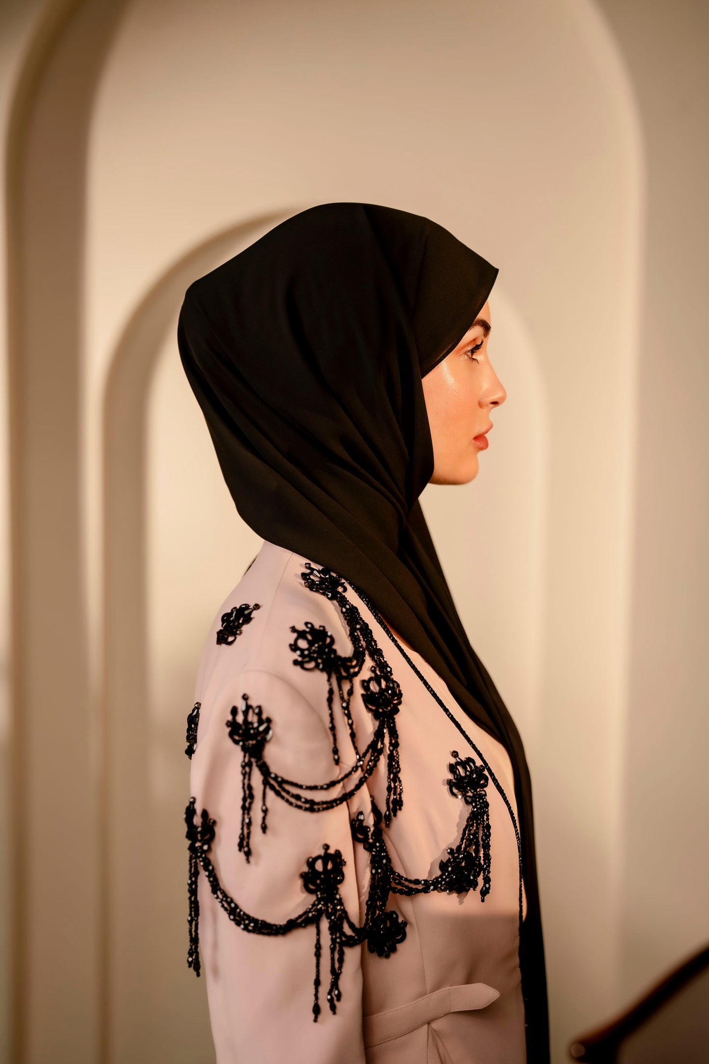 Woman wearing a black hijab and a white outfit with black embellishments in a neutral setting.
