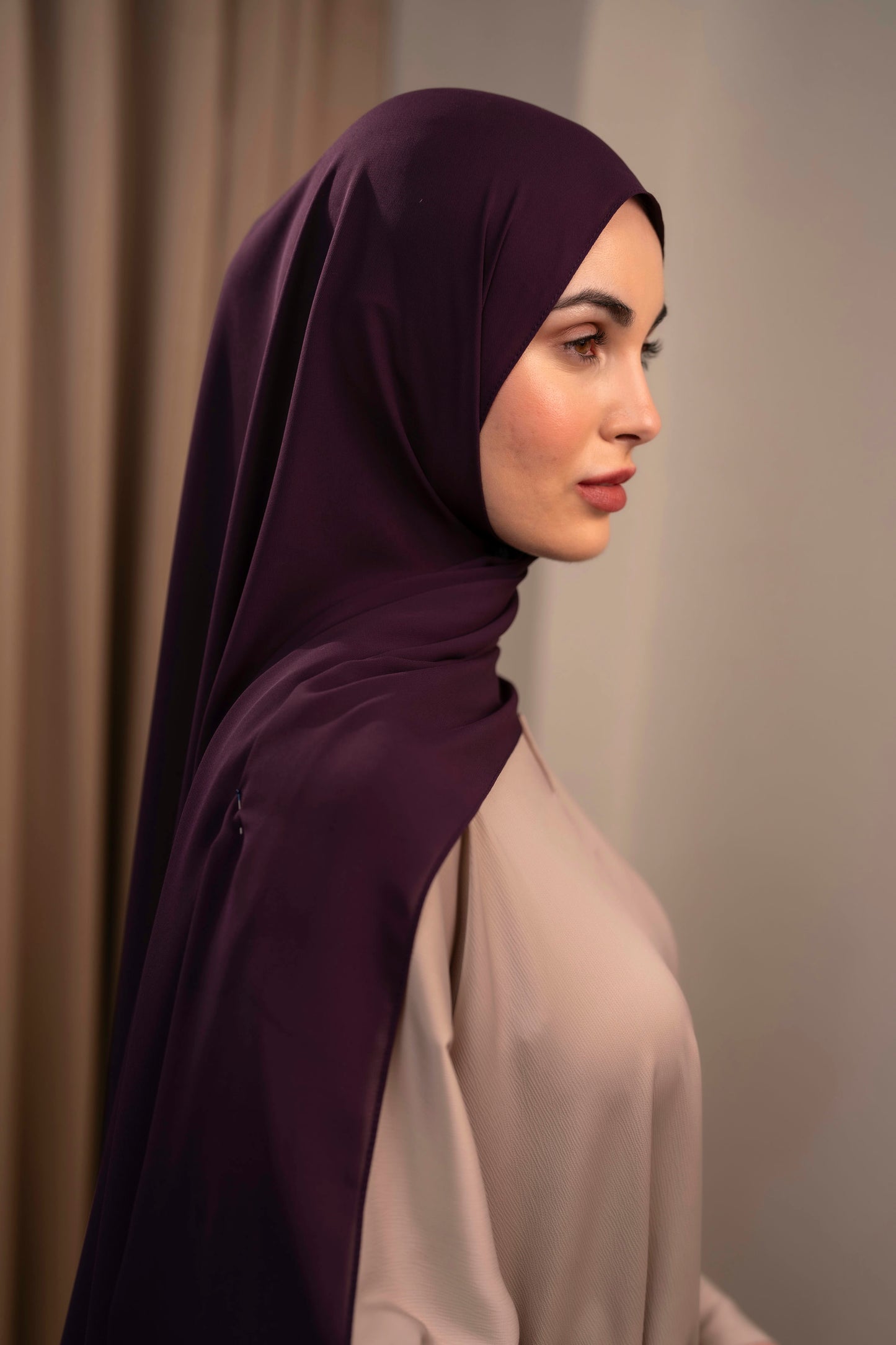 Woman wearing a dark purple hijab and beige top against a neutral background - Plum