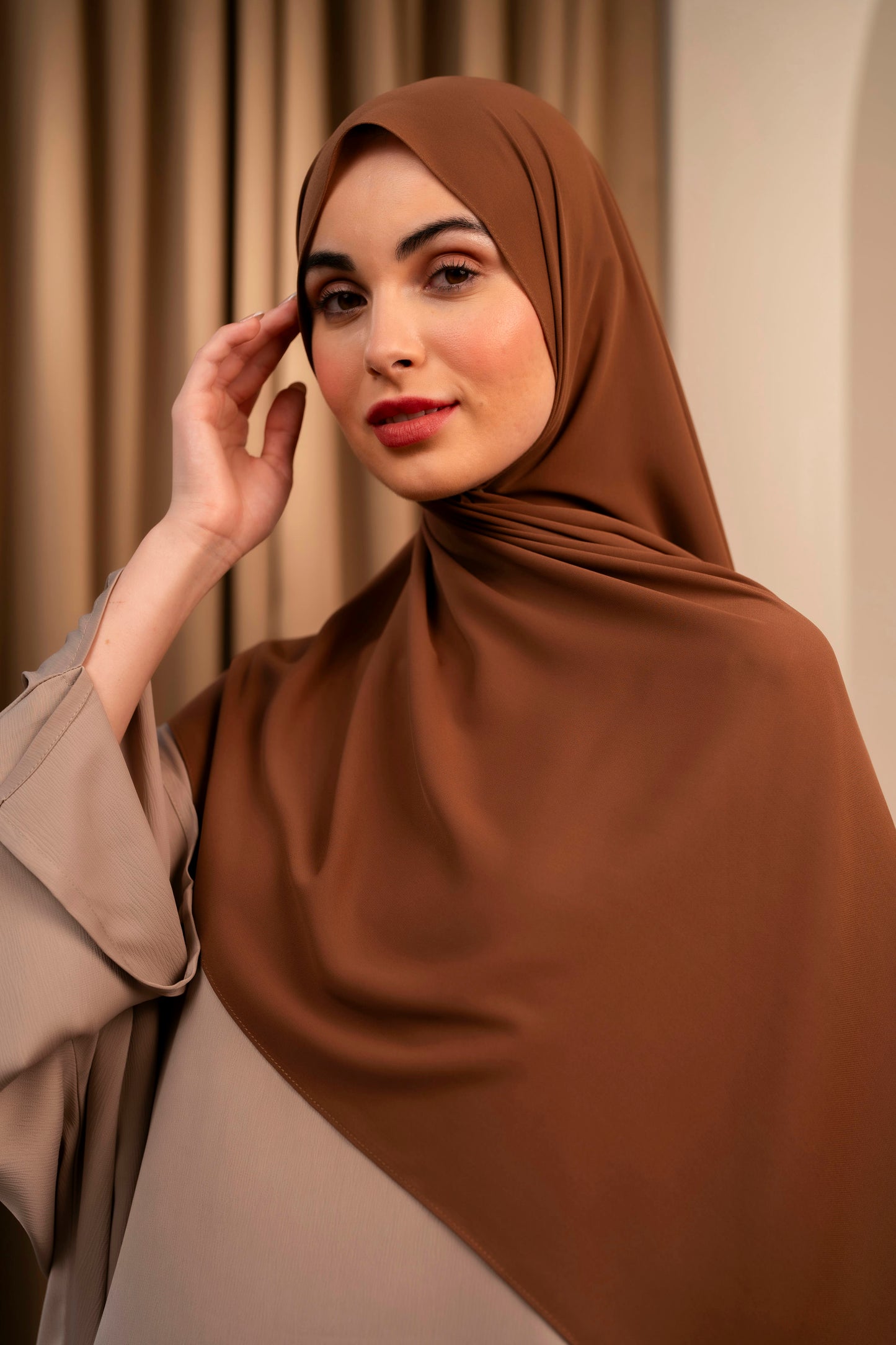 Woman wearing a brown hijab against a beige curtain background