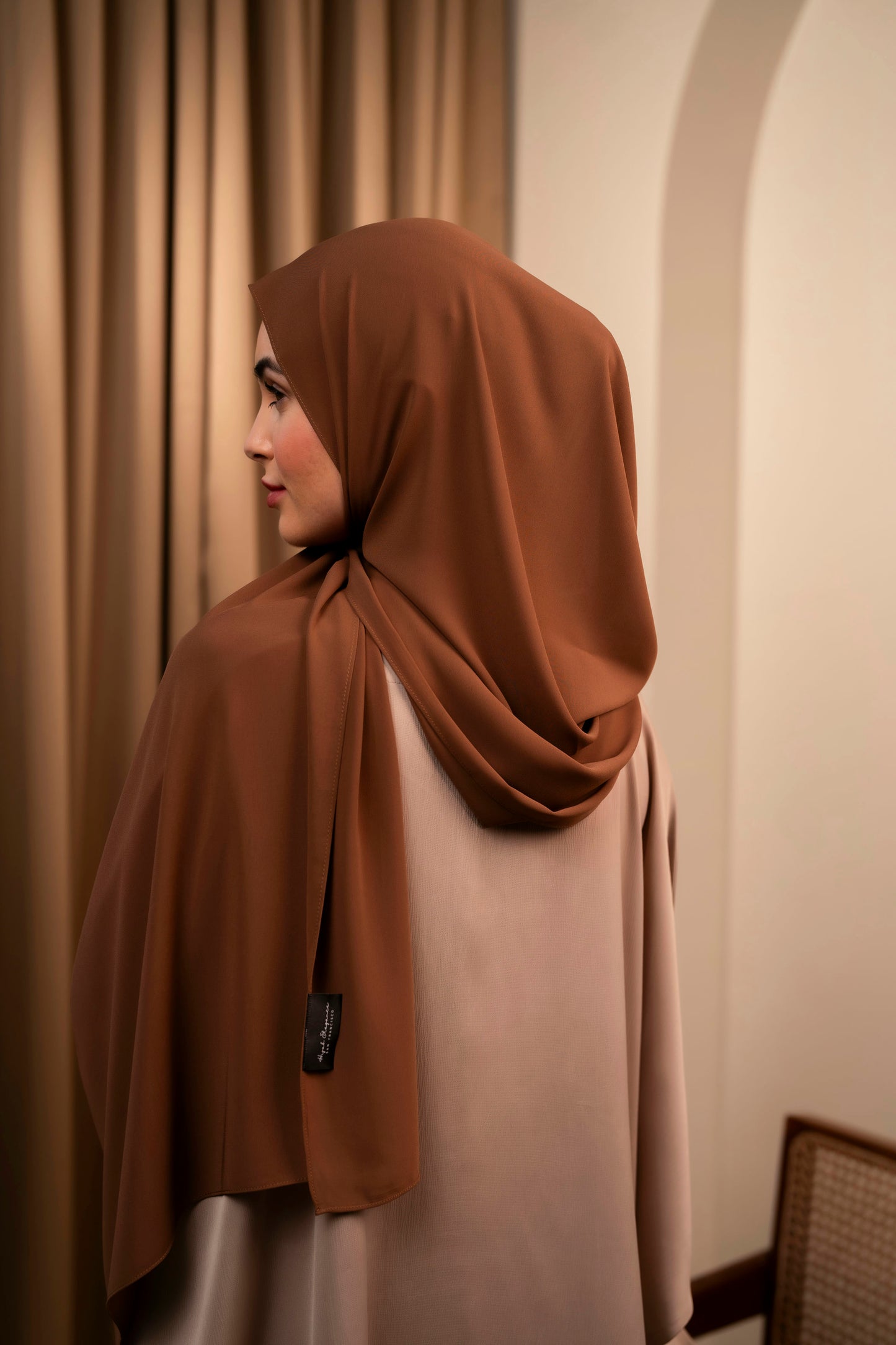 Woman wearing a brown hijab and white dress standing against a beige wall.