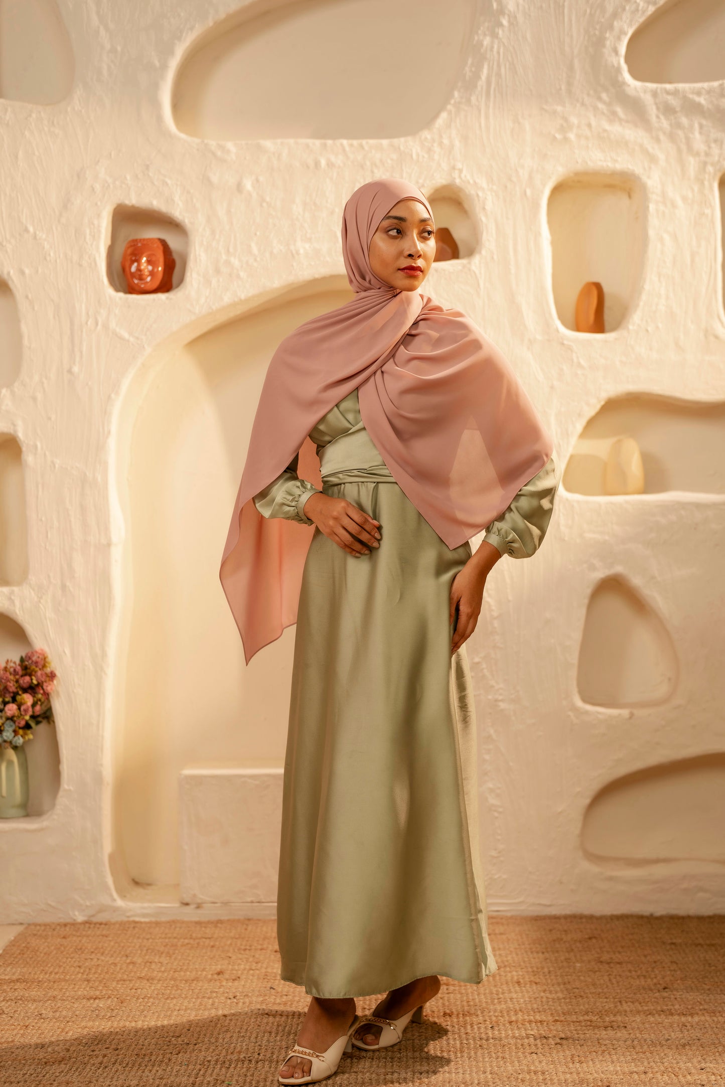 Woman in a light green dress and pink hijab standing in a modern, softly lit room with abstract wall designs - dusty pink