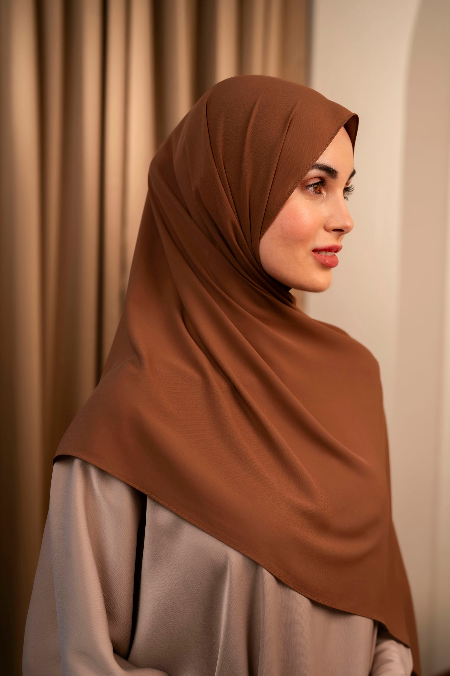Woman wearing a brown hijab against a beige curtain background