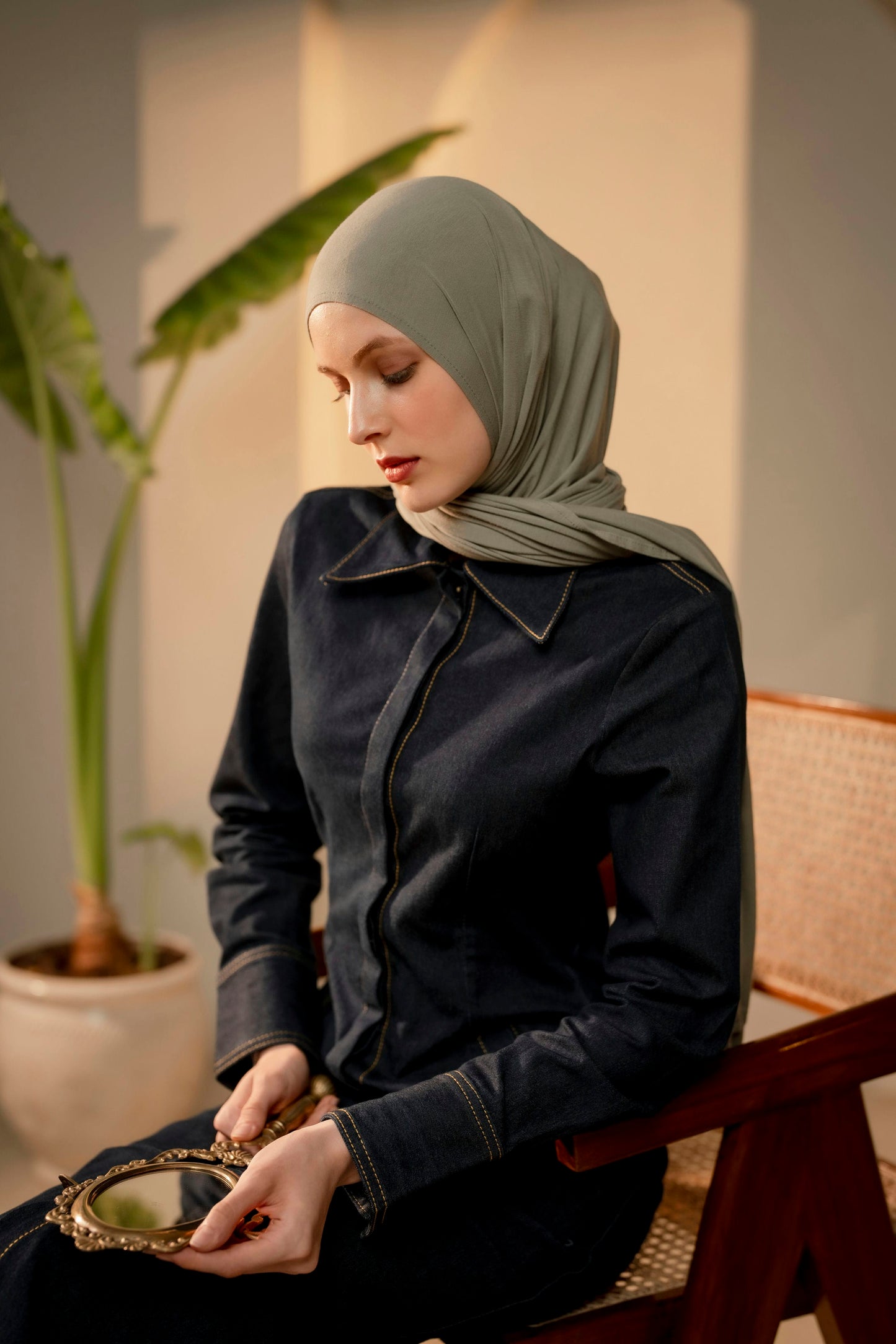 Woman wearing a sea green hijab and dark jacket sitting indoors with a plant in the background. She is holding a small antique mirror