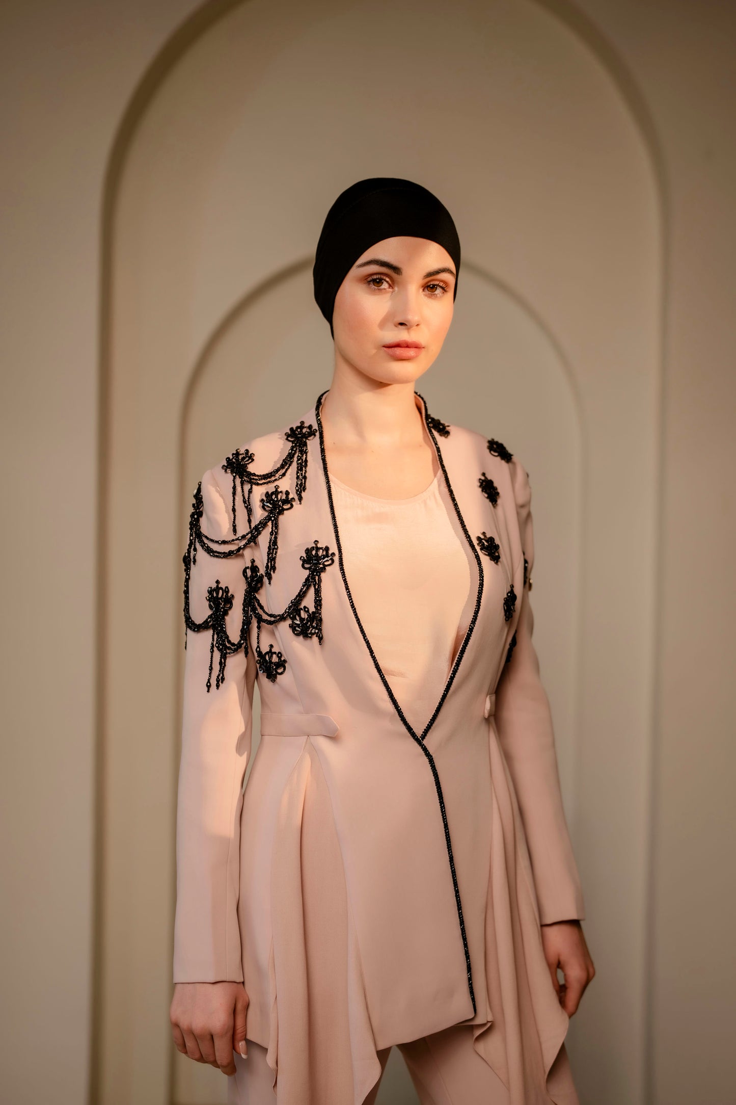 Woman wearing a beige outfit with black embroidery against a neutral background