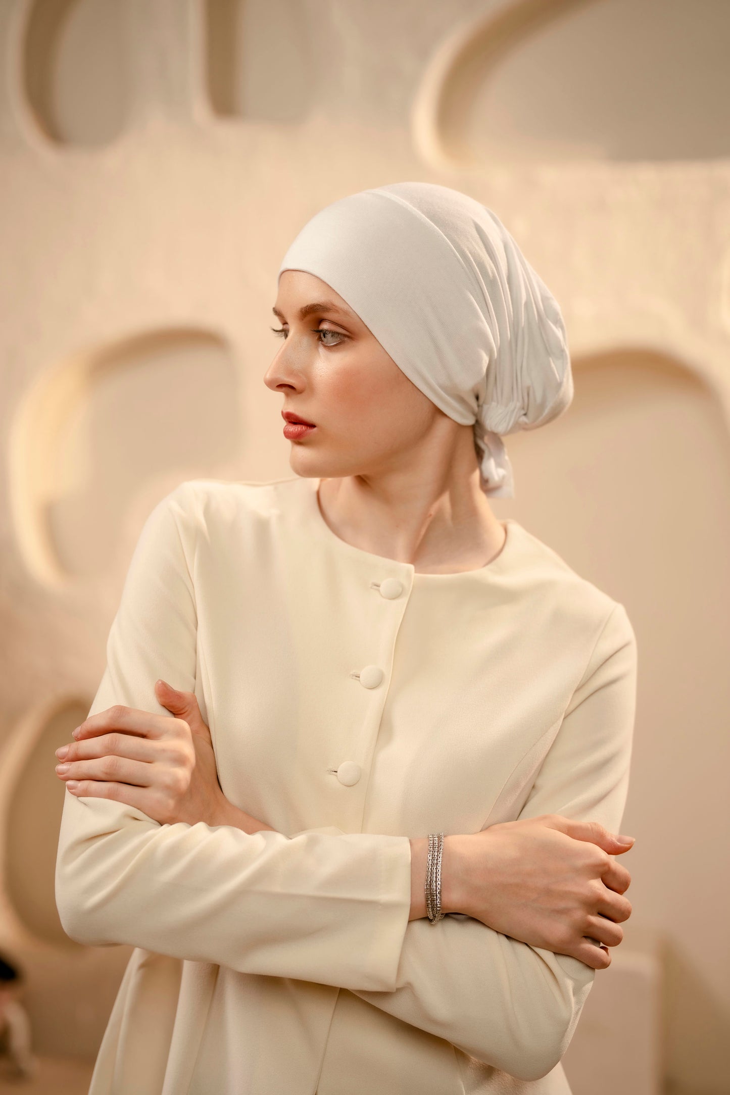 Woman wearing a white headscarf and outfit against a beige wall.