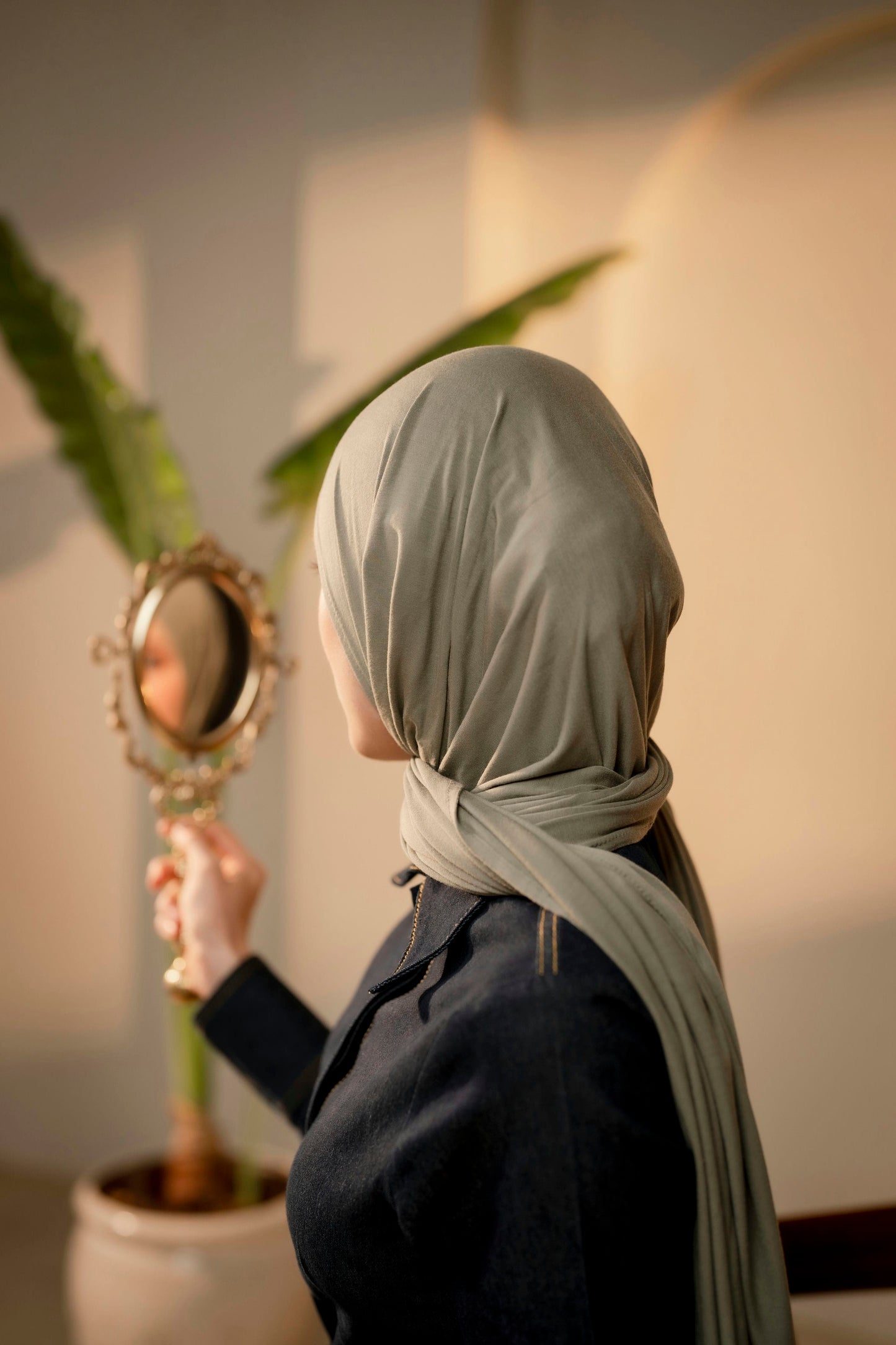 Person wearing a light green hijab holding a small mirror indoors.