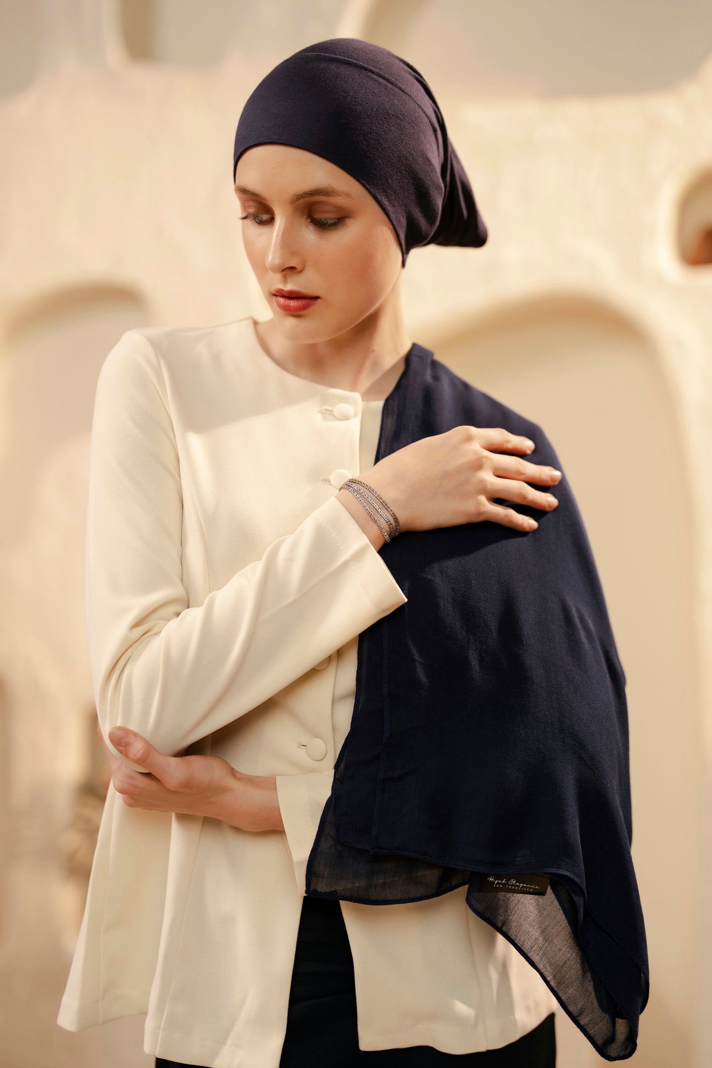 Woman wearing a white coat and navy headscarf against a beige textured background