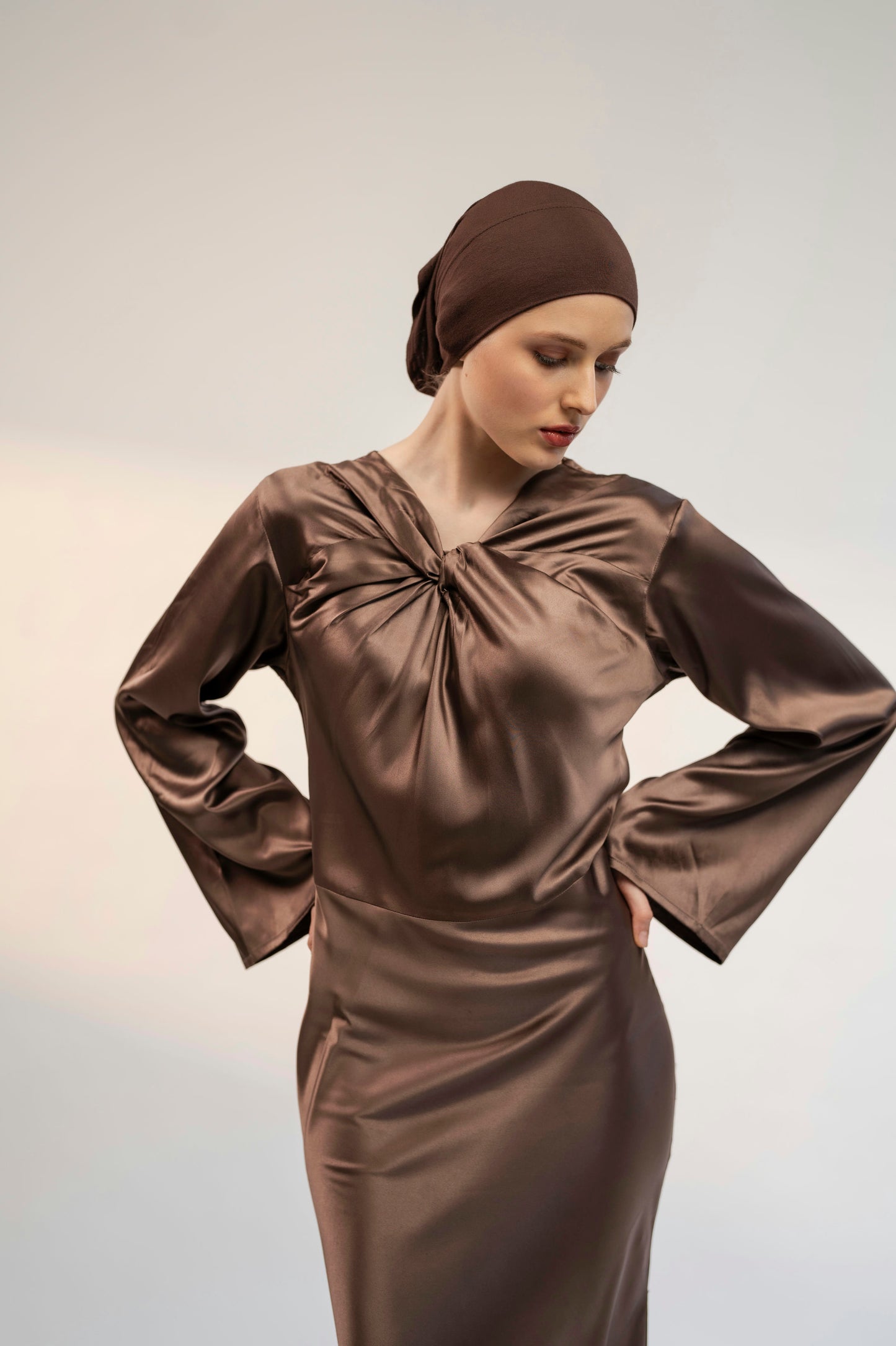 Woman wearing a coffee brown satin dress and headscarf against a plain background