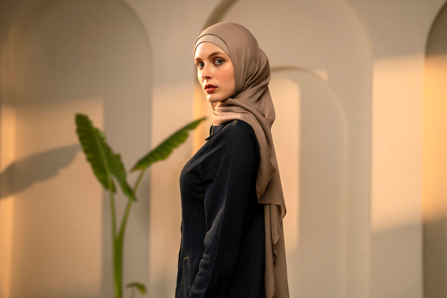 Woman wearing a beige hijab and dark clothing in an archway with a plant - mocha