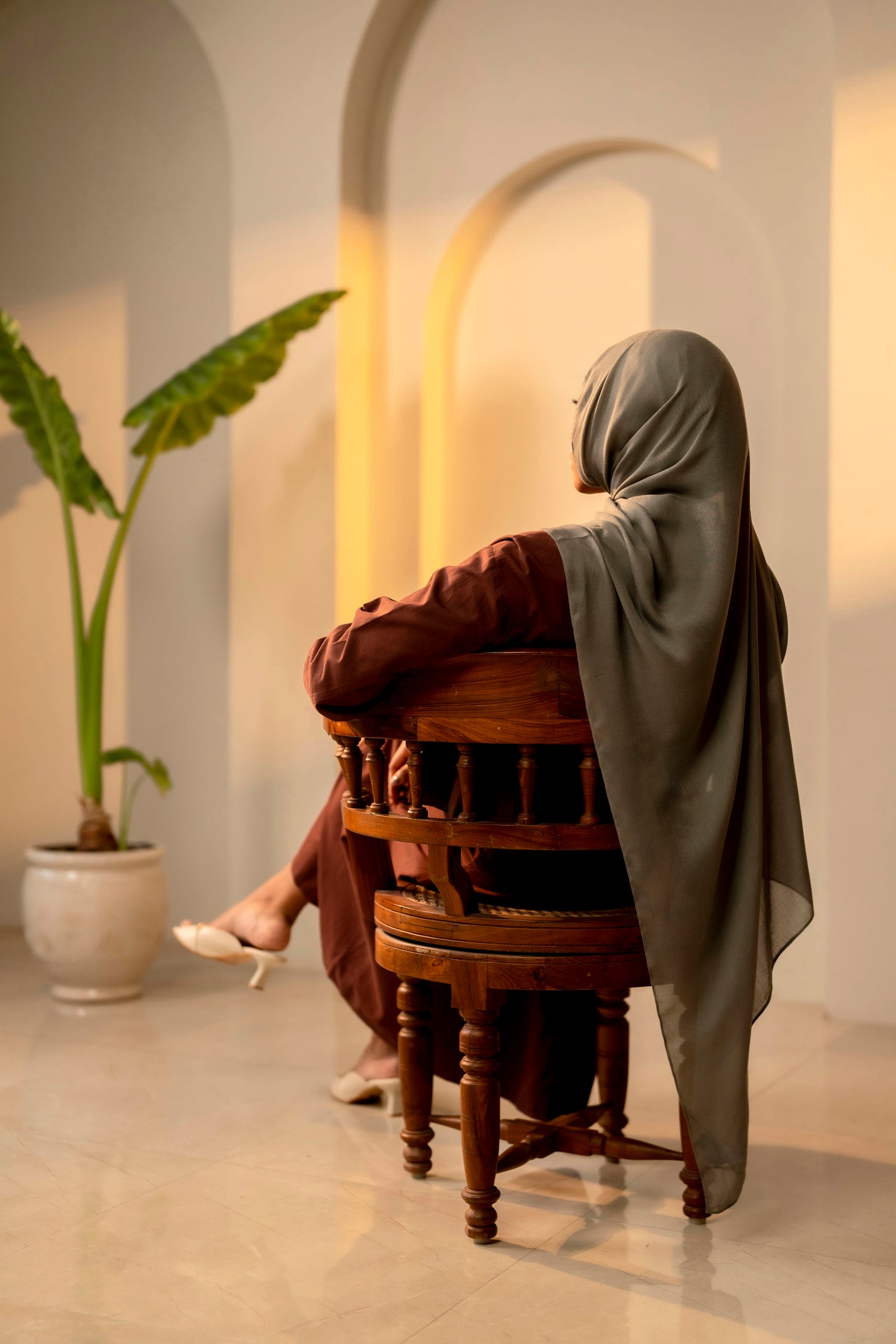 Person sitting on a wooden chair in a softly lit room with a plant in the background - sage green