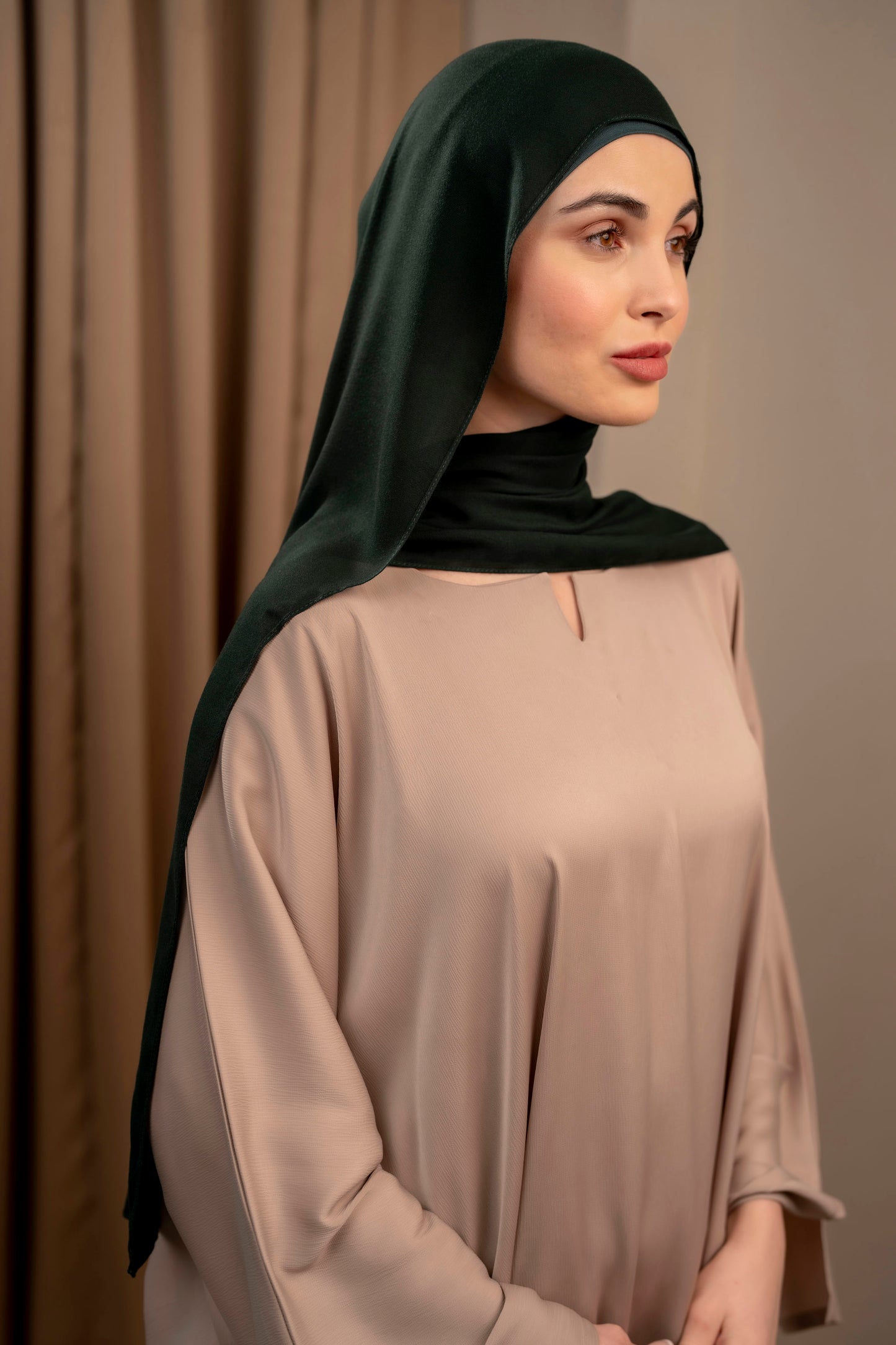 Woman wearing a forest green hijab and beige outfit against a plain background - forest green