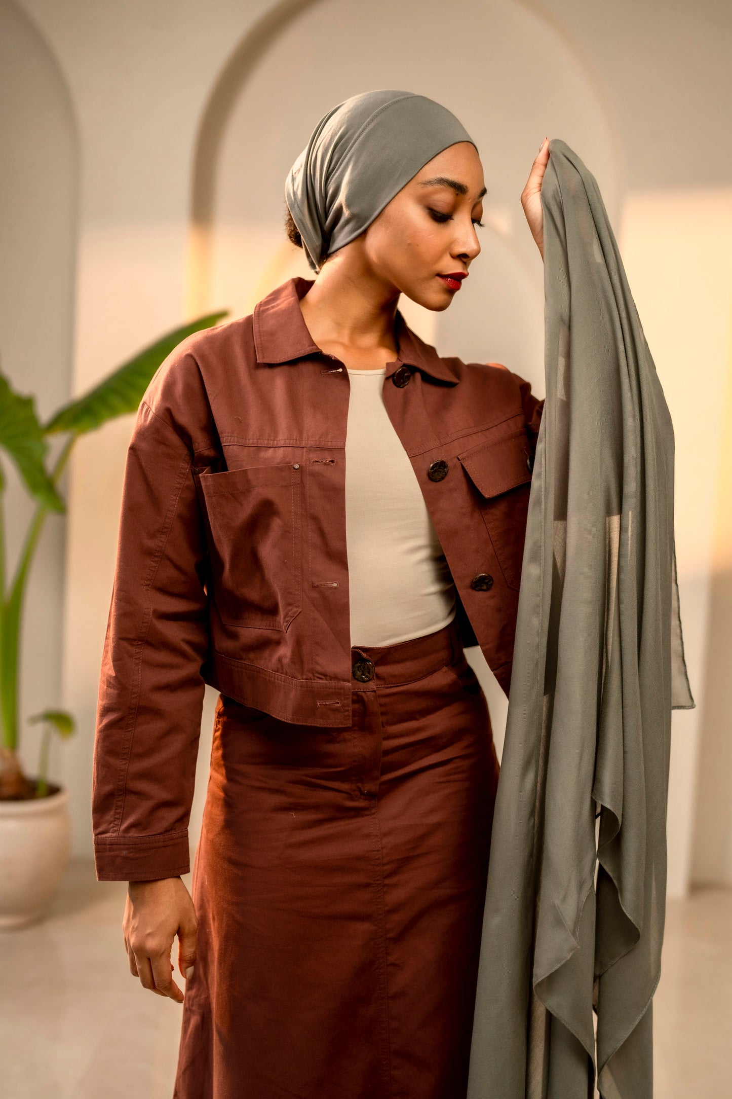 Woman in a brown jacket and skirt holding a gray coat in a neutral setting - sage green