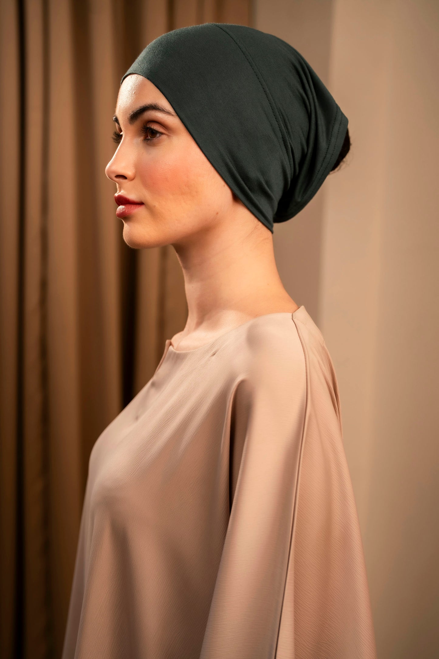 Woman wearing a forest green headscarf and beige top against a neutral background