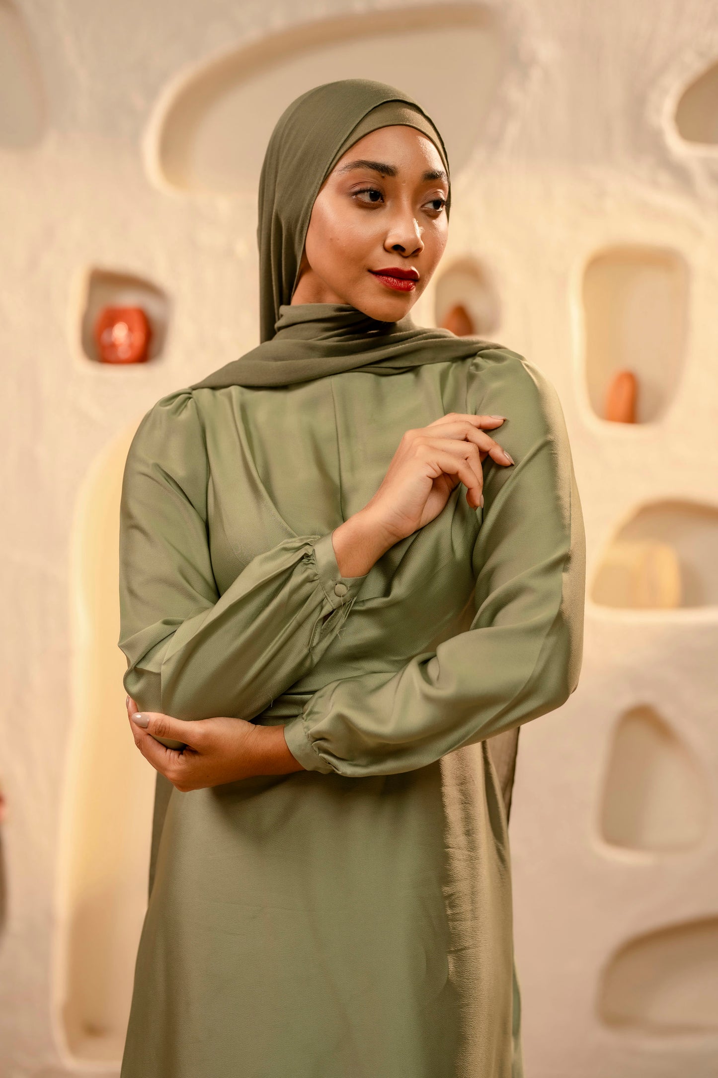 Woman wearing a olive green hijab and dress against an abstract wall.