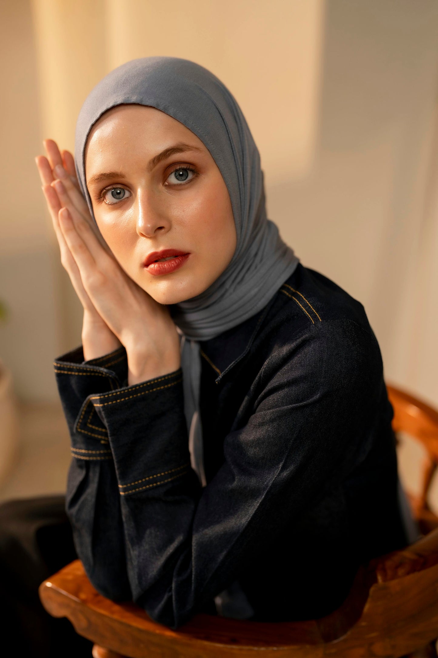 Woman wearing a light gray hijab in an indoor setting sitting on a wooden chair hands raised till the face and face leaning on the hands