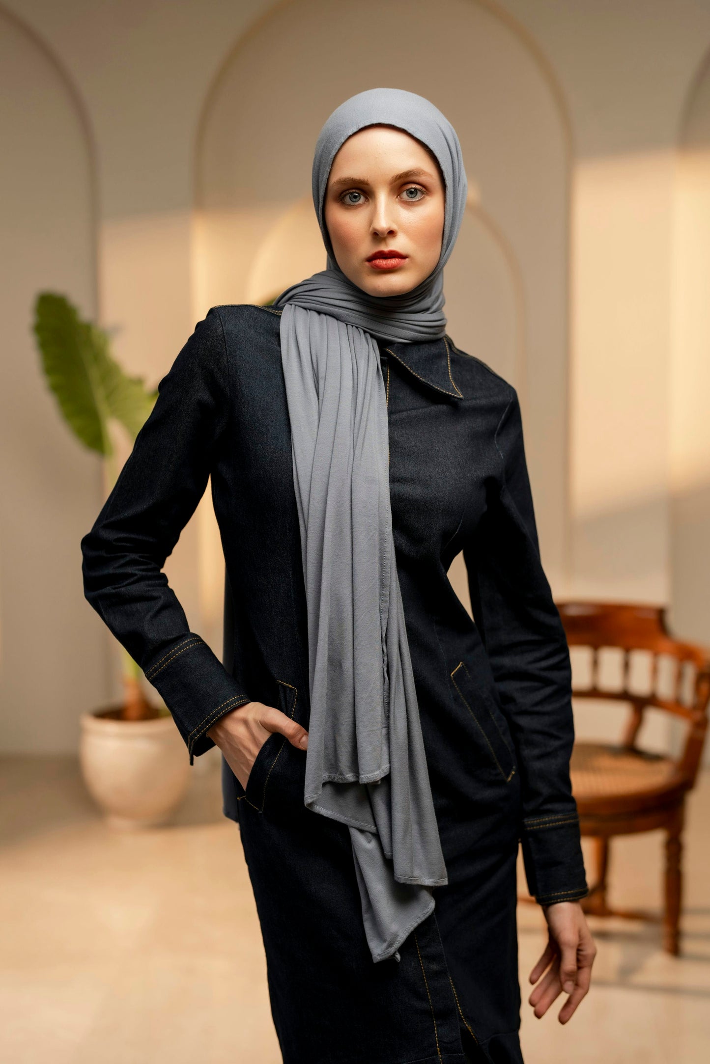 Woman front facing wearing a light gray hijab in an indoor setting with arches and a plant.