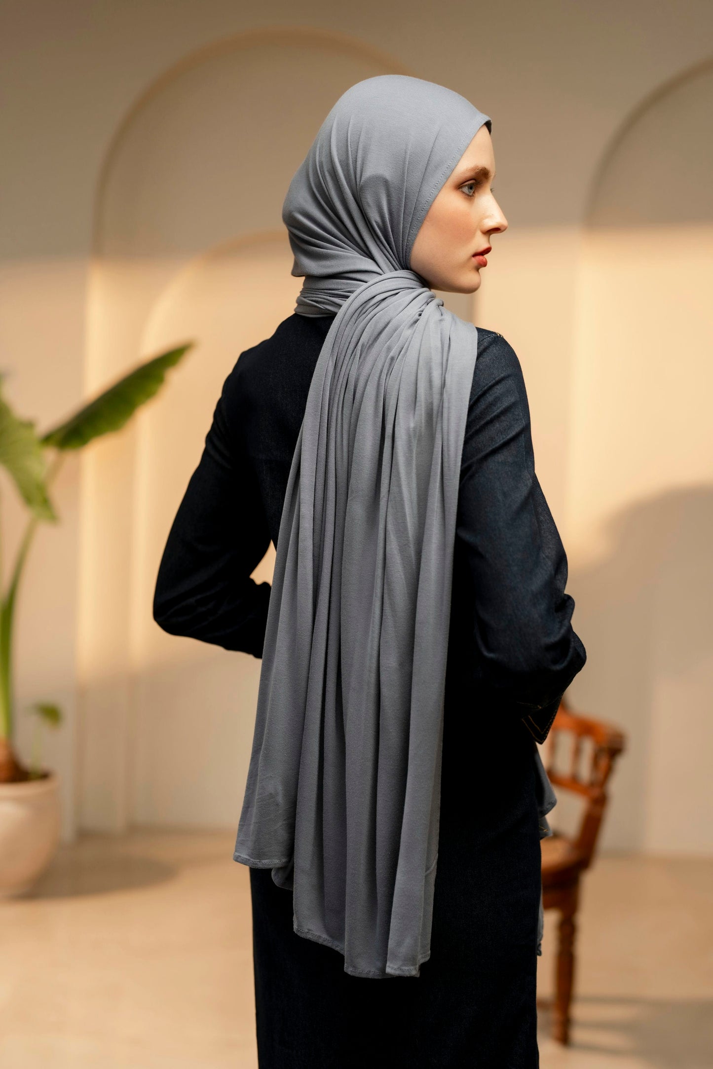 Woman wearing a light gray hijab in an indoor setting with arches and a plant.