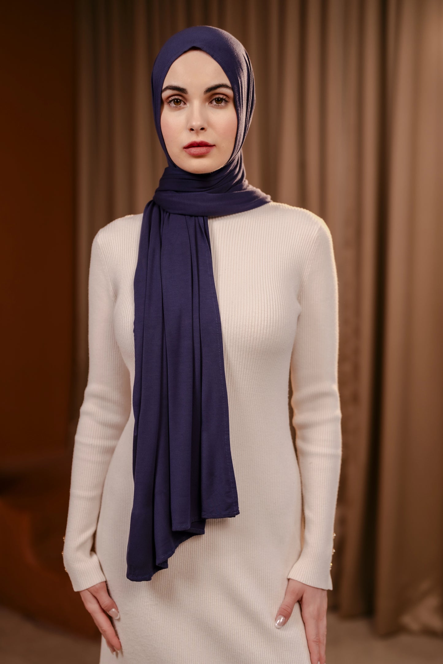 Woman wearing a navy hijab and white outfit against a brown curtain background