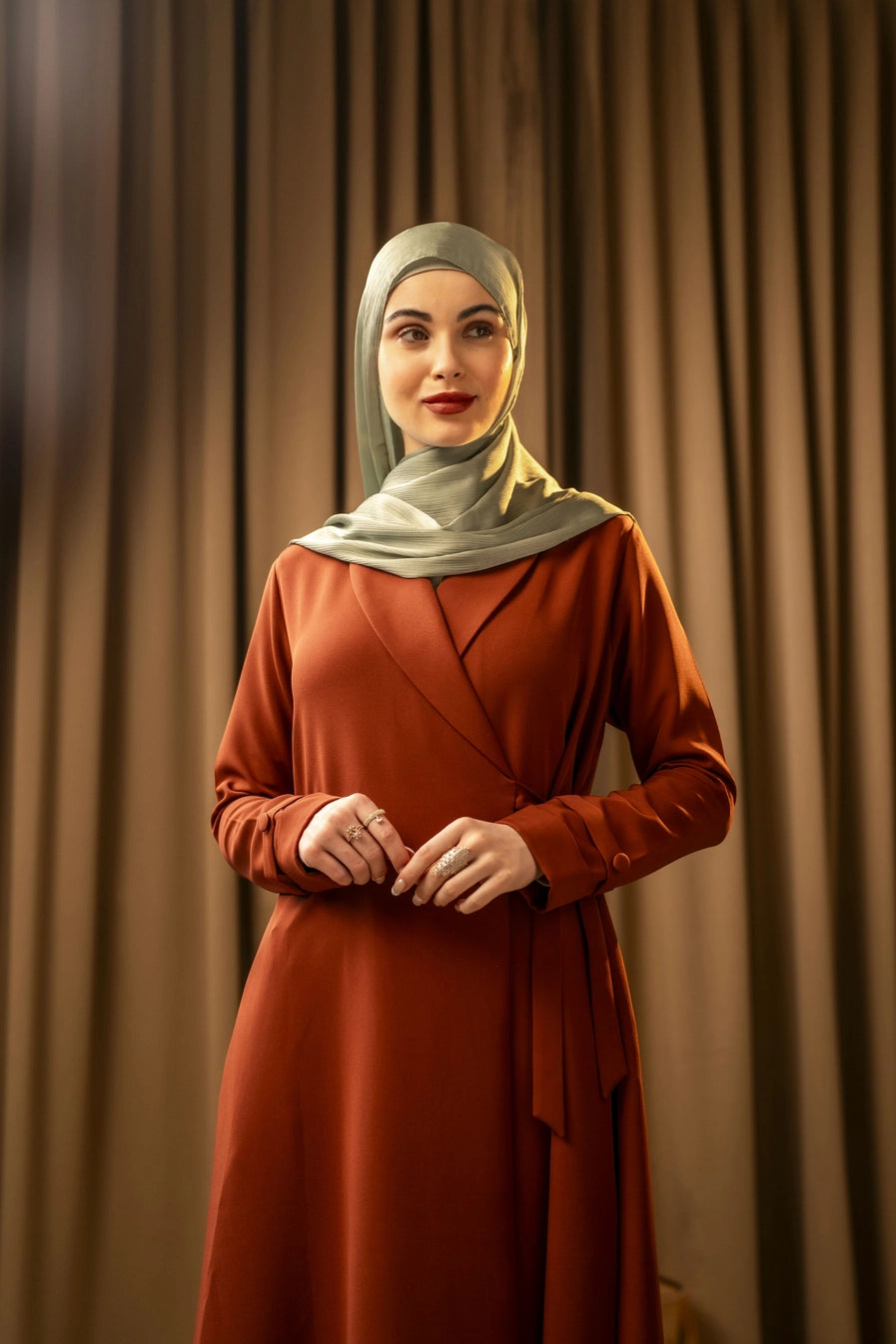 Woman wearing a rust-colored dress and light green hijab against a brown curtain background