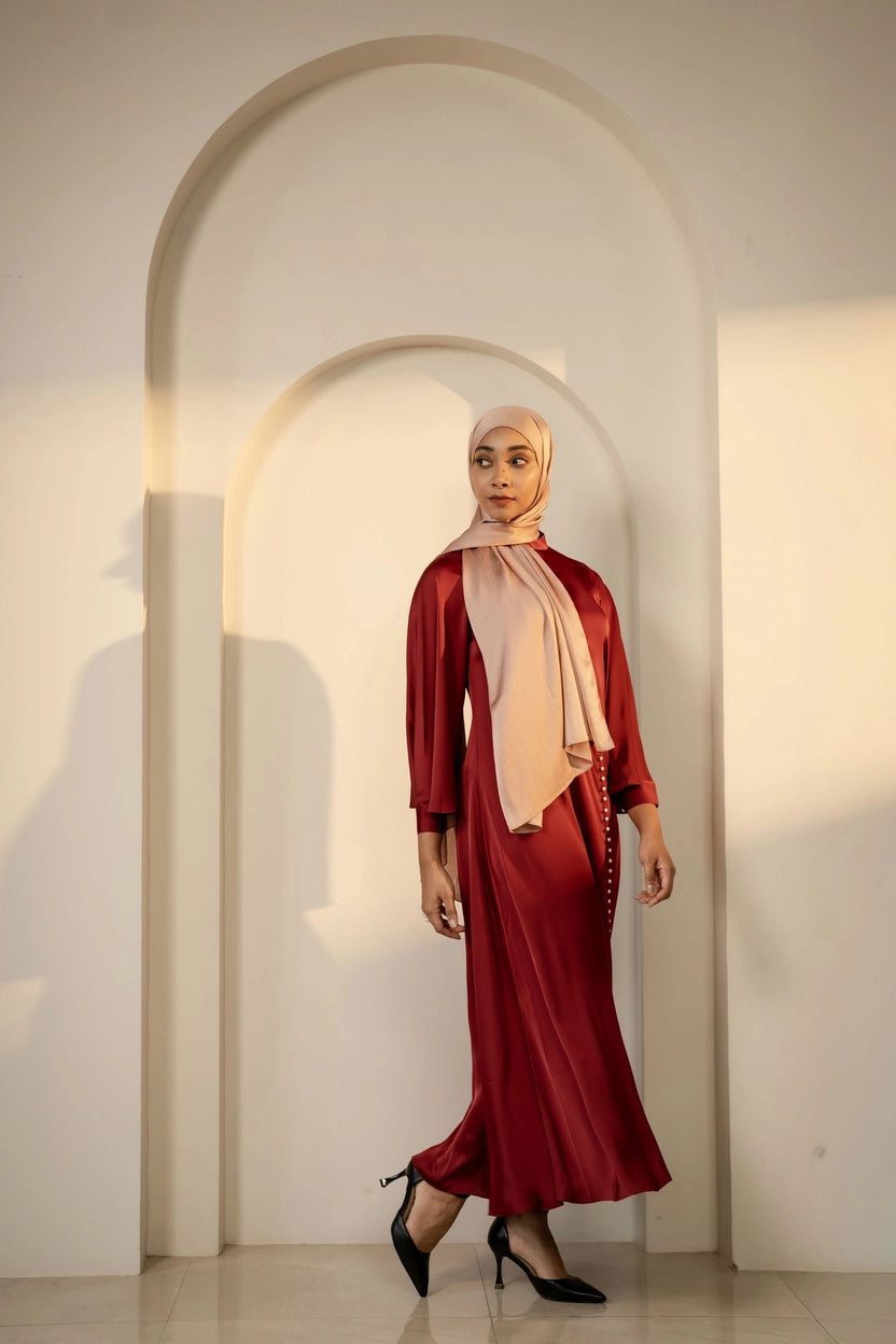 Woman in a maroon outfit with a beige copper hijab standing in front of an archway.