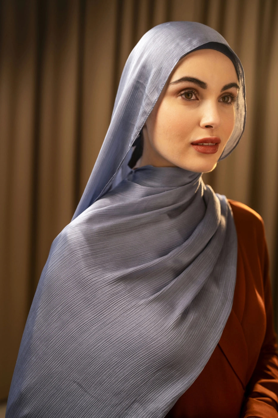 Woman wearing a light blue hijab against a beige curtain background