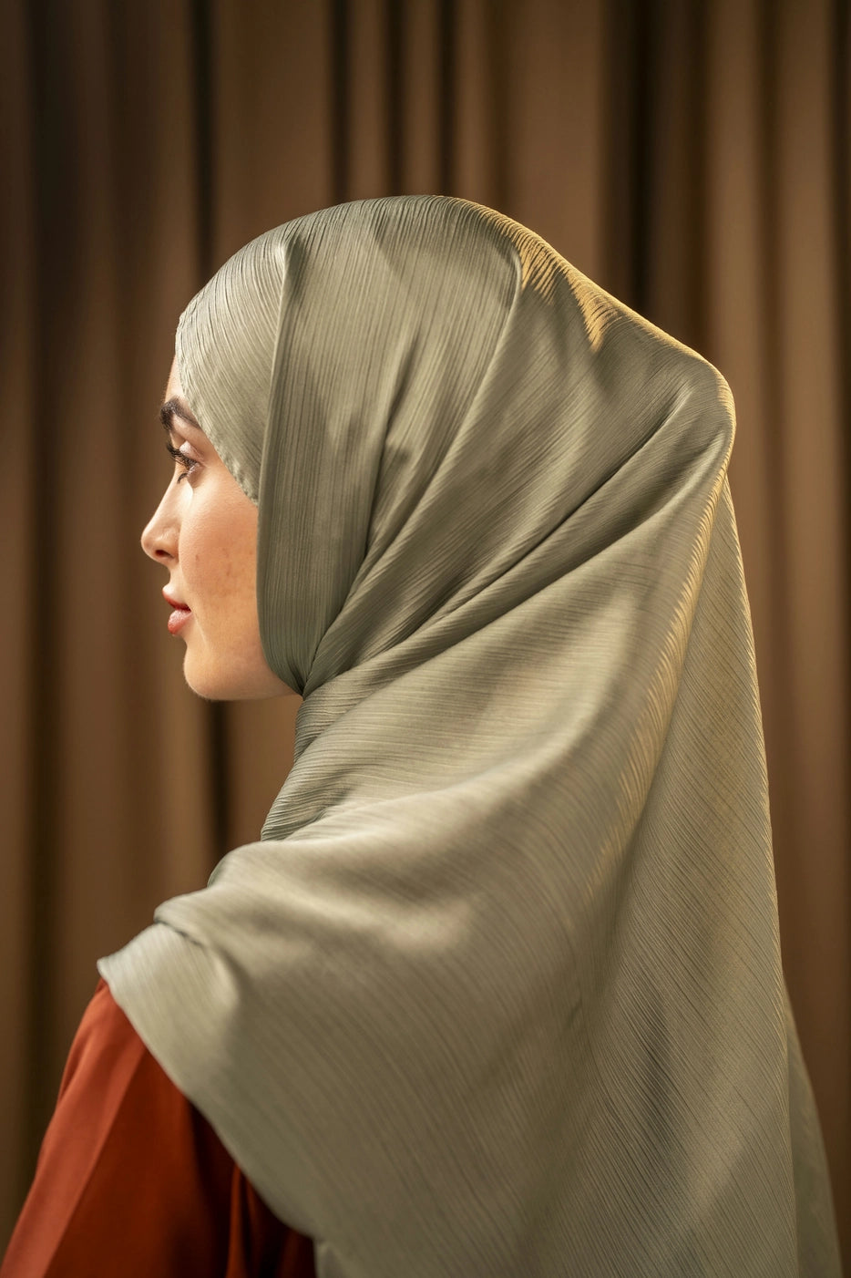 Woman wearing a light green hijab against a brown curtain background
