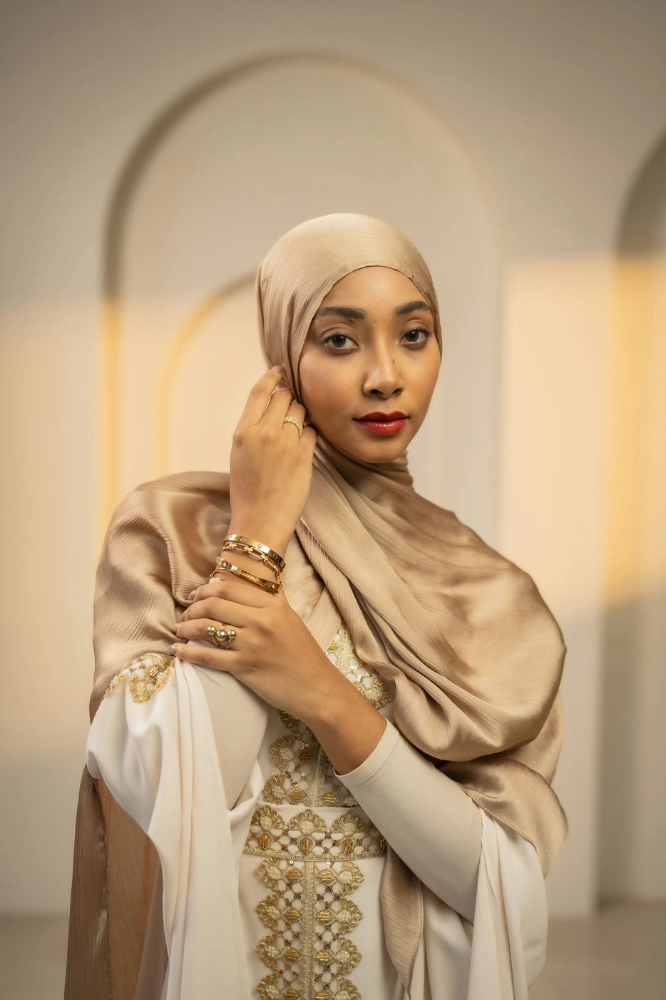 Woman wearing a beige gold hijab and white dress with gold embroidery in an indoor setting.