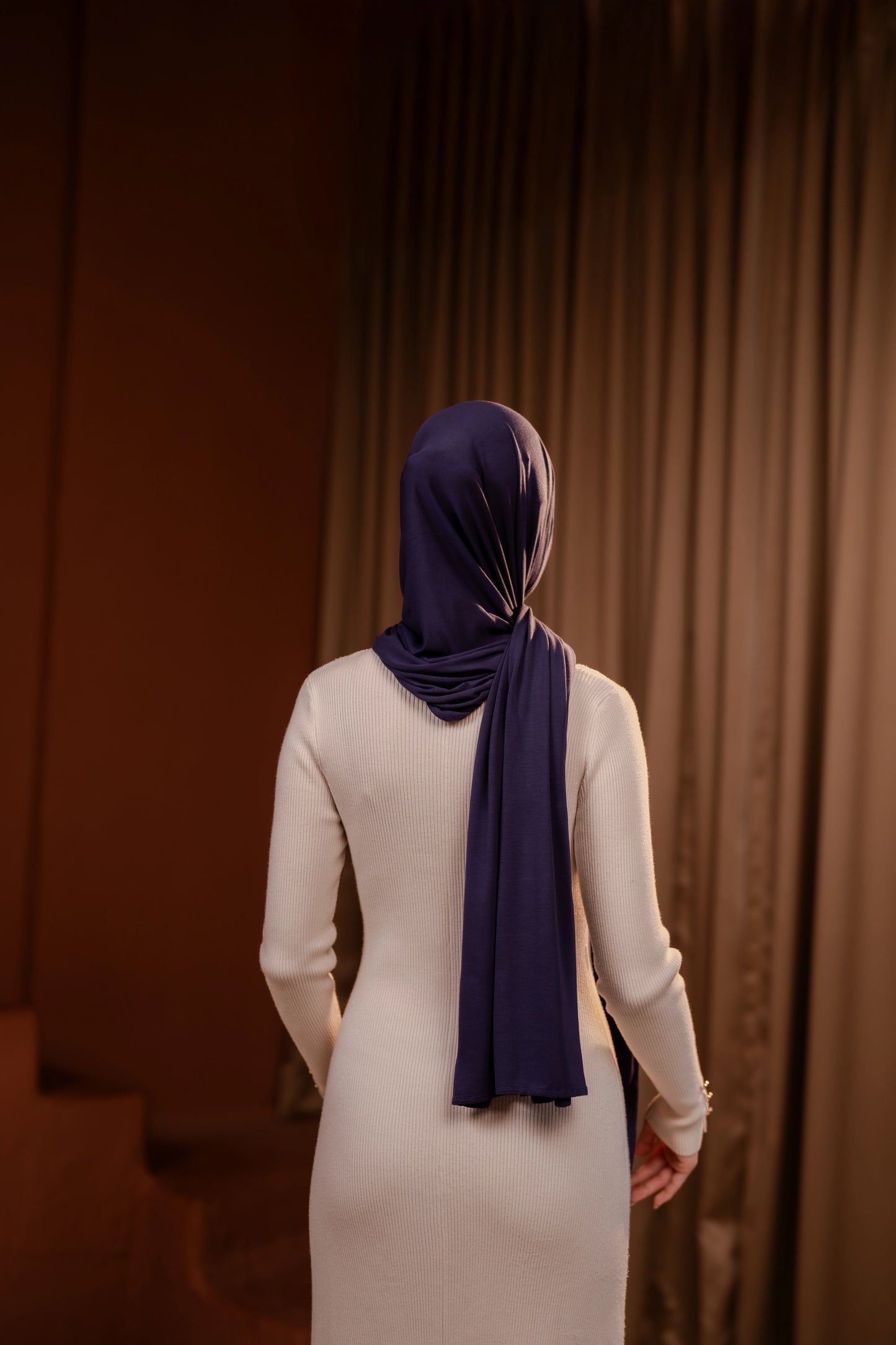 Person wearing a white dress and purple hijab standing in front of a brown curtain.