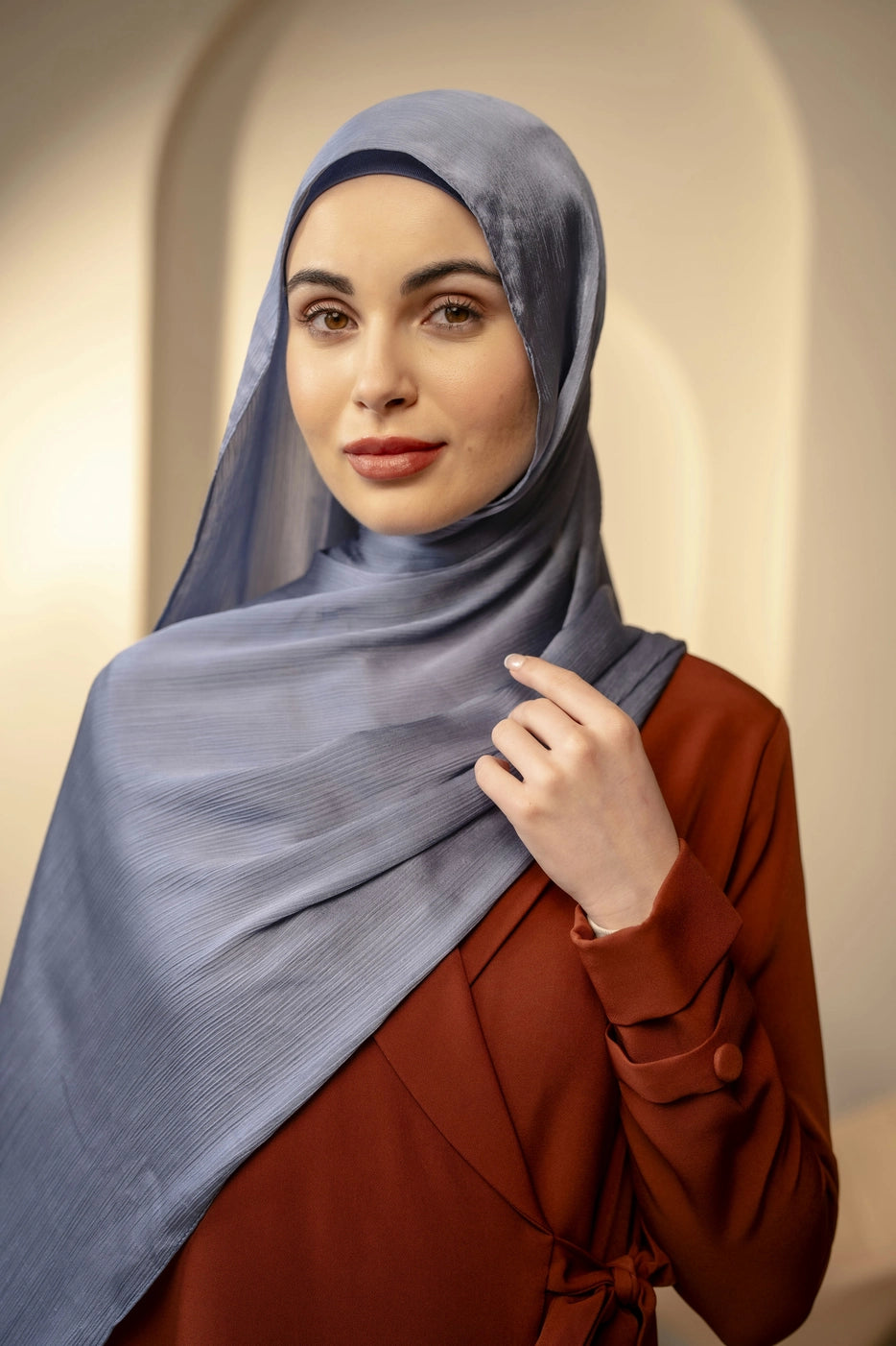 Woman wearing a sky blue hijab and brown outfit against a beige background