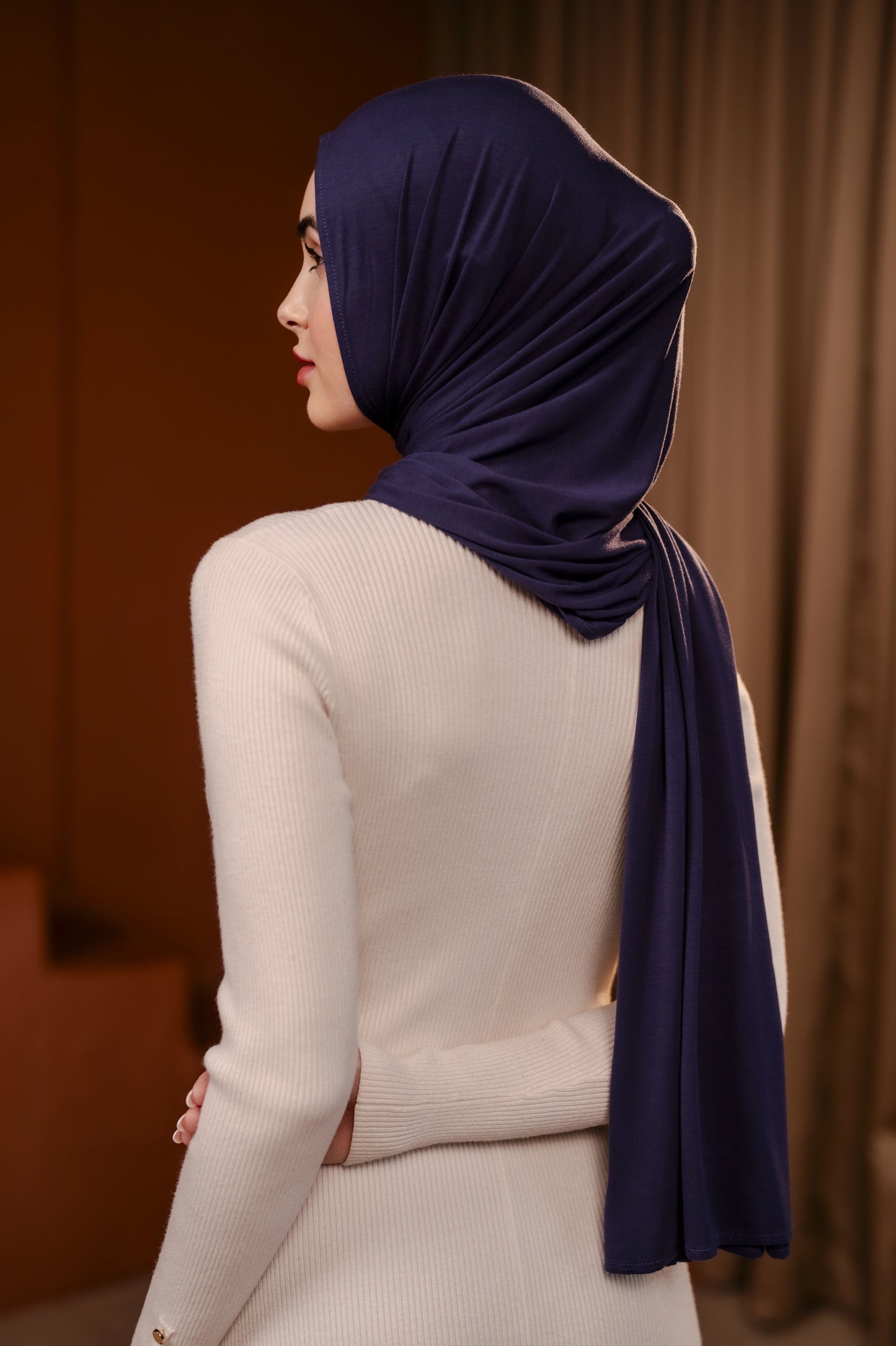 Woman wearing a dark blue hijab and beige outfit against a brown background