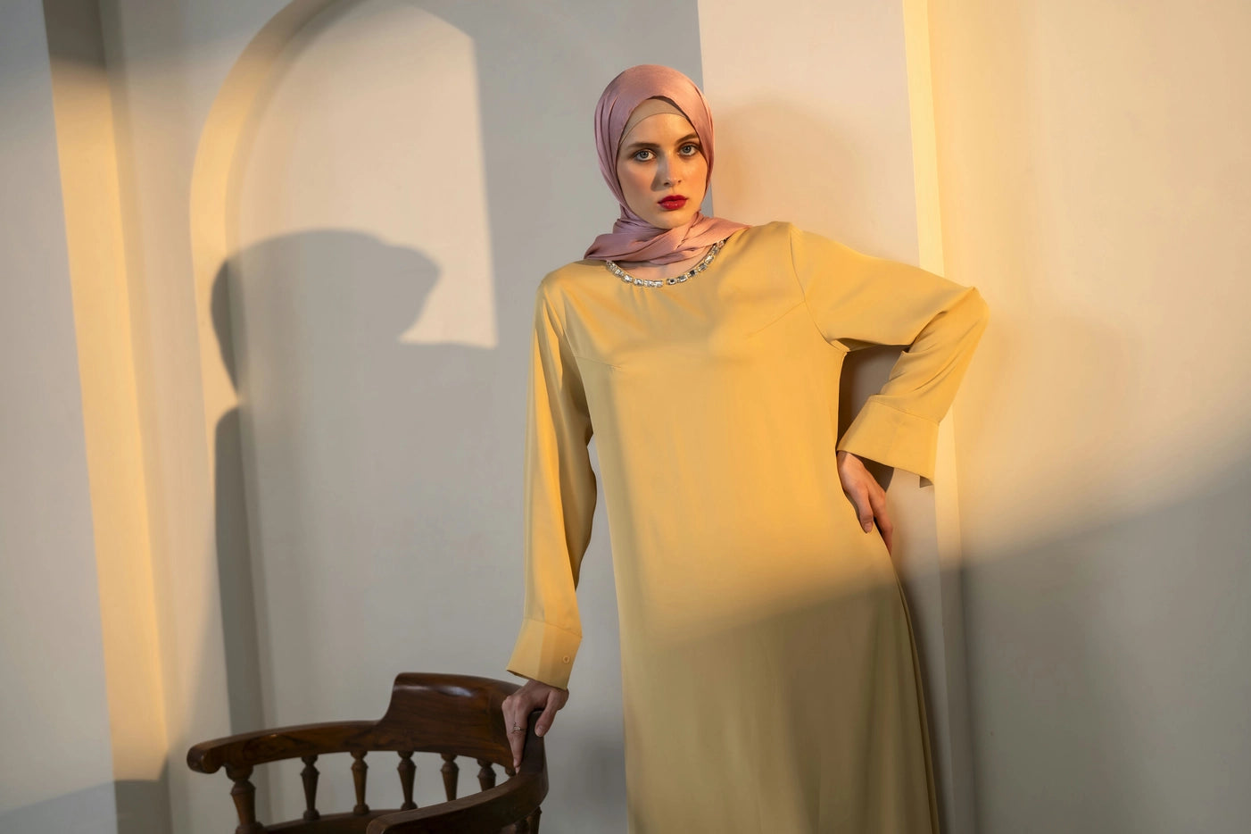 Woman in a yellow dress and pink hijab standing in a softly lit room.