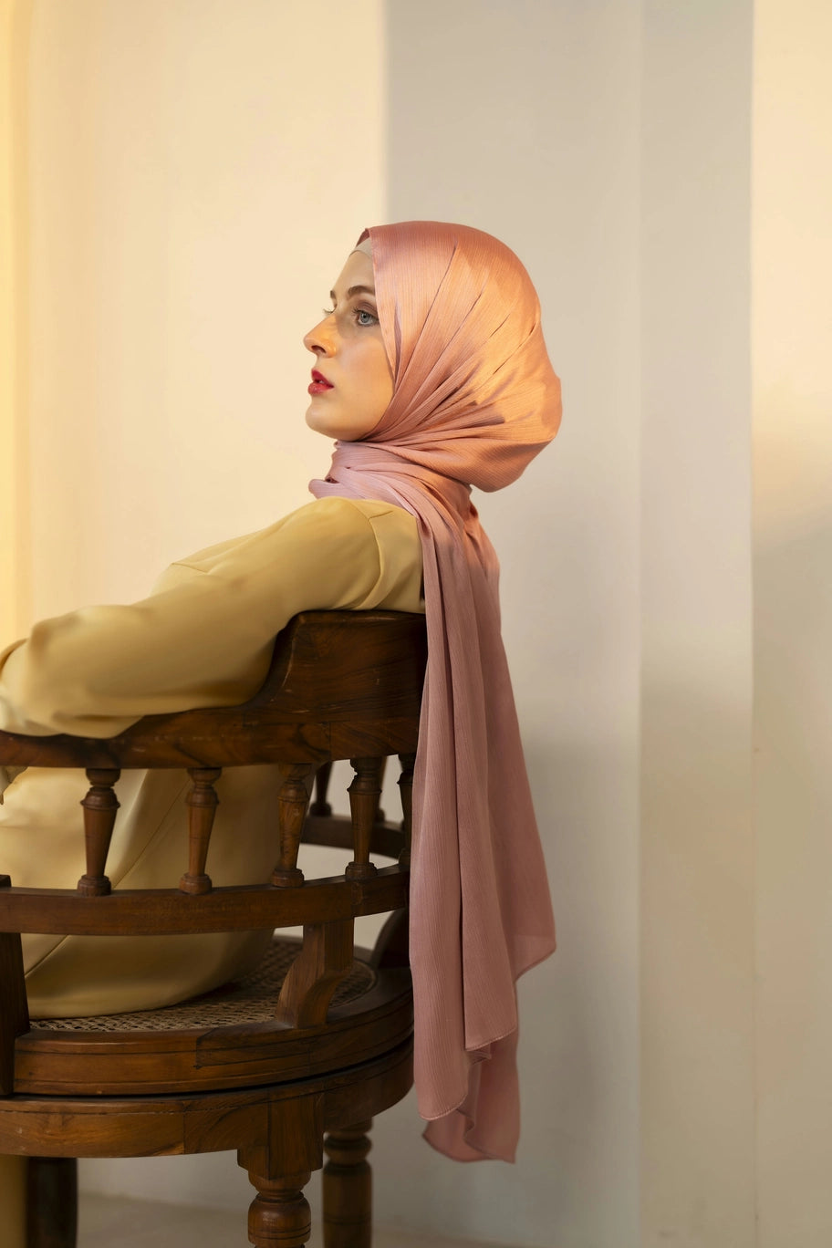 Woman wearing a pink hijab sitting on a wooden chair against a light-colored wall.