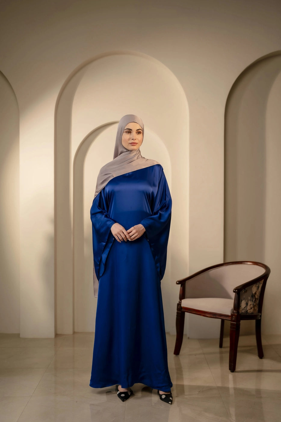 Woman in a dark navy dress with silver gray hijab standing in an archway with a chair in the background