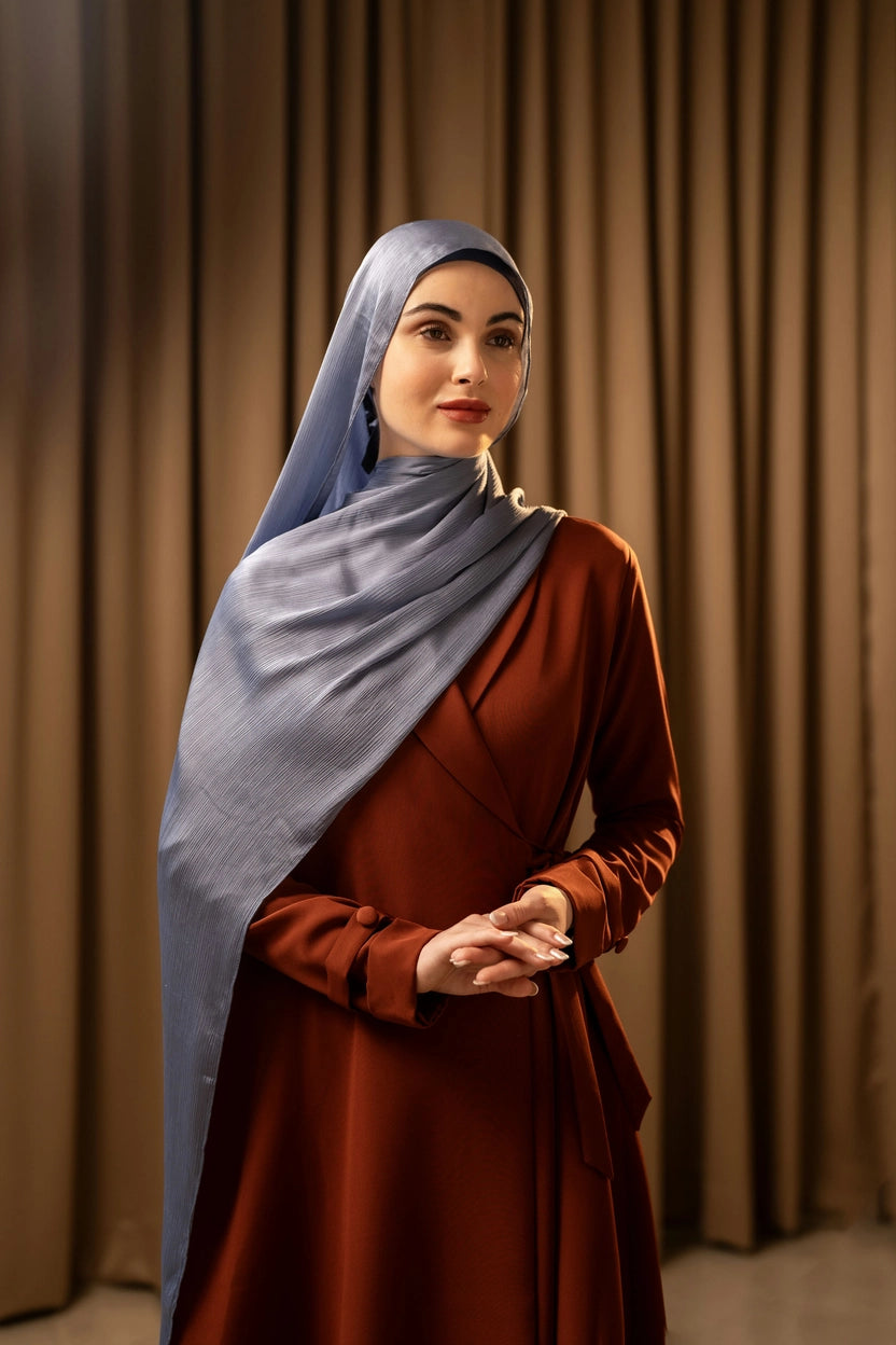 Woman wearing a blue hijab and brown dress against a beige curtain background