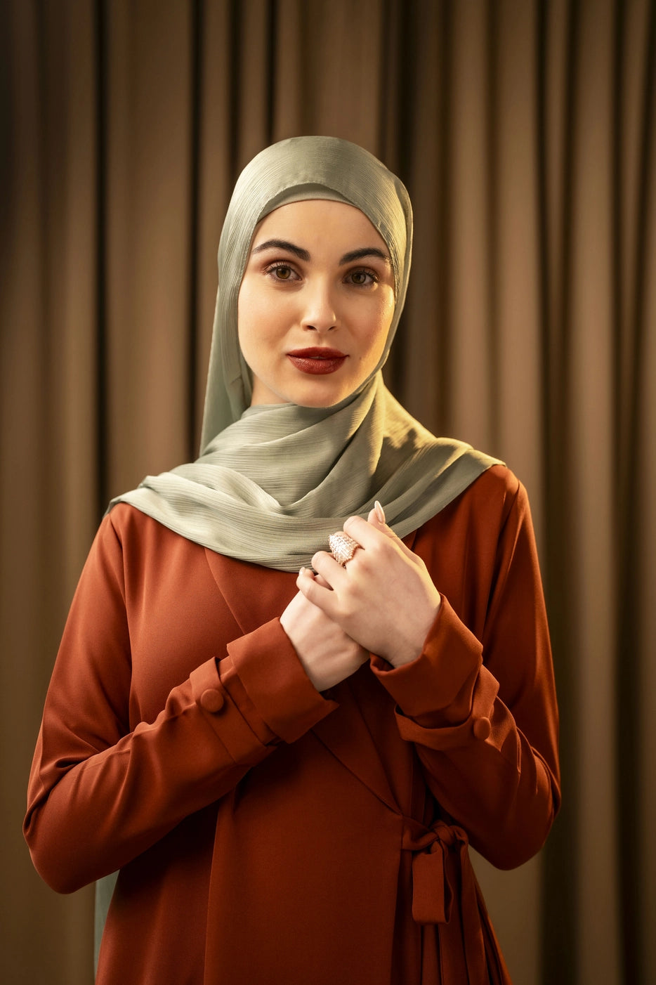 Woman wearing a green hijab and brown outfit against a brown curtain background