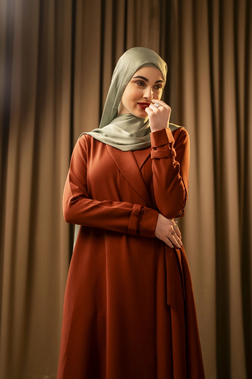 Woman wearing a rust-colored dress and luxury light green hijab against a beige curtain background