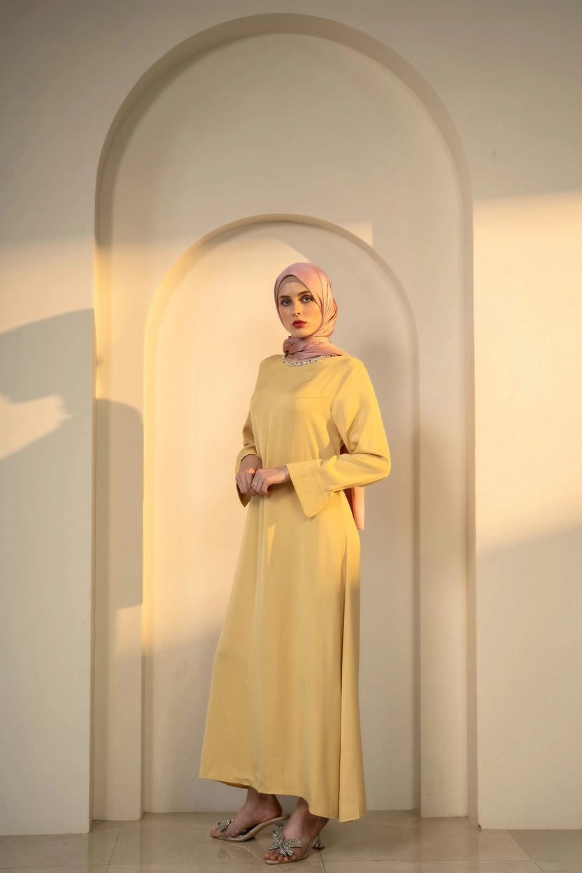 Woman in a yellow dress and pink hijab standing in an archway with warm lighting
