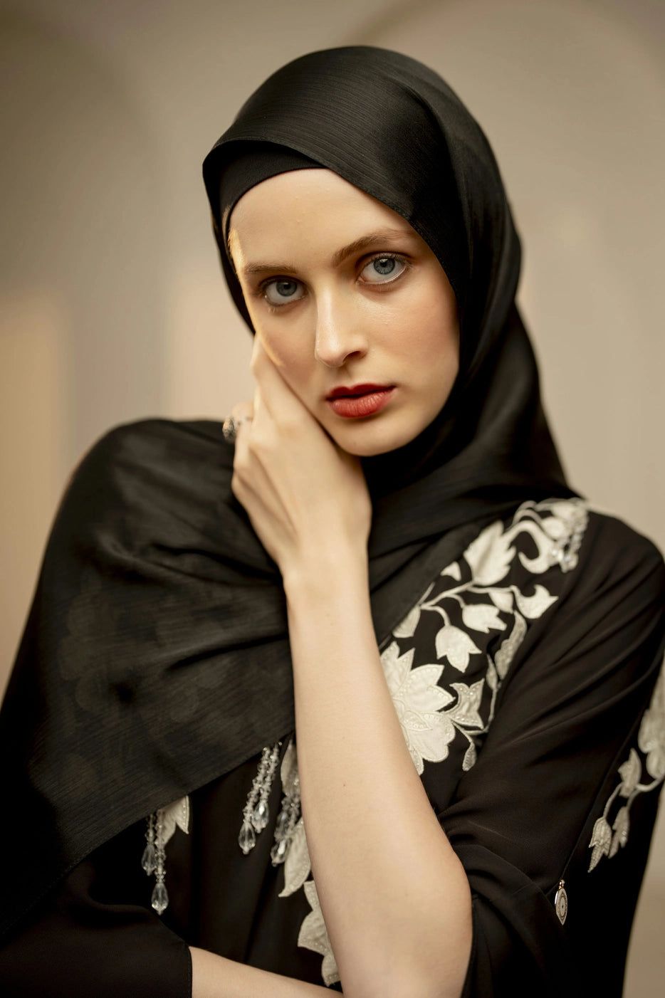 Woman wearing a black hijab with white embroidery, posing against a neutral background