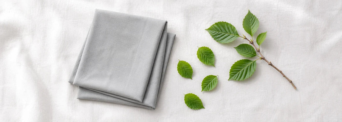 Folded grey modal hijab laid next to fresh beech leaves on white linen — illustrating modal fabric's sustainable beech wood pulp origin — Hijab Elegance