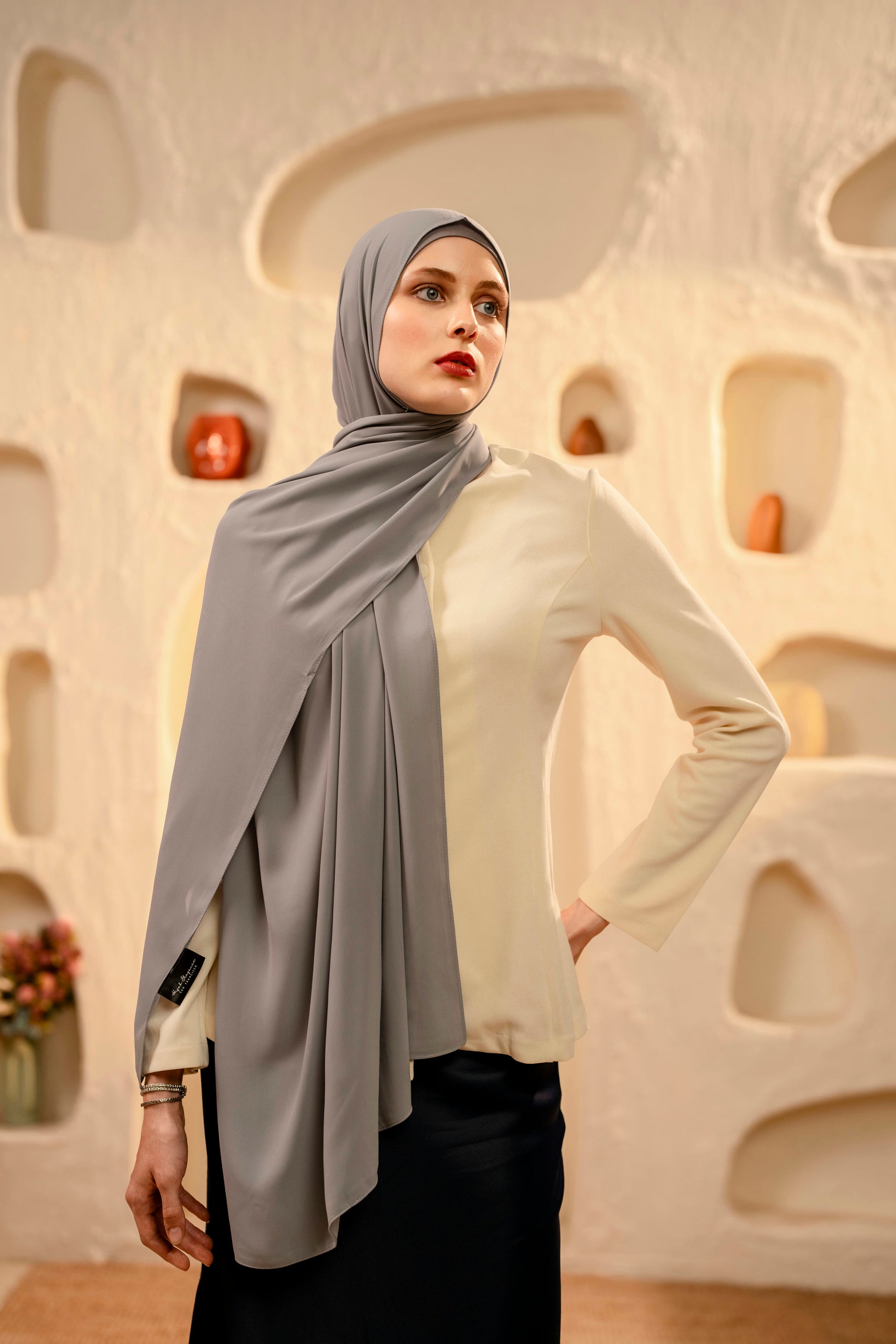 Woman wearing a gray hijab and white top against an abstract wall.