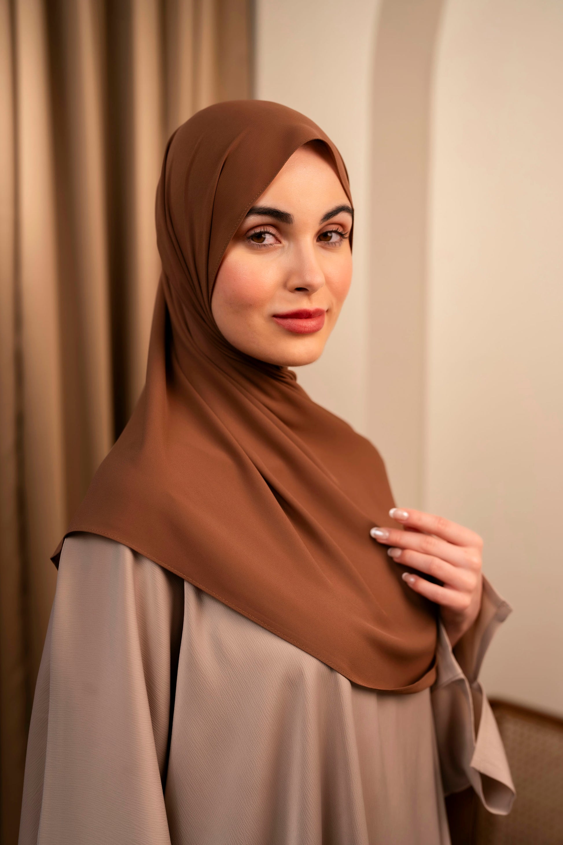 Woman wearing a brown hijab and beige outfit against a beige curtain background