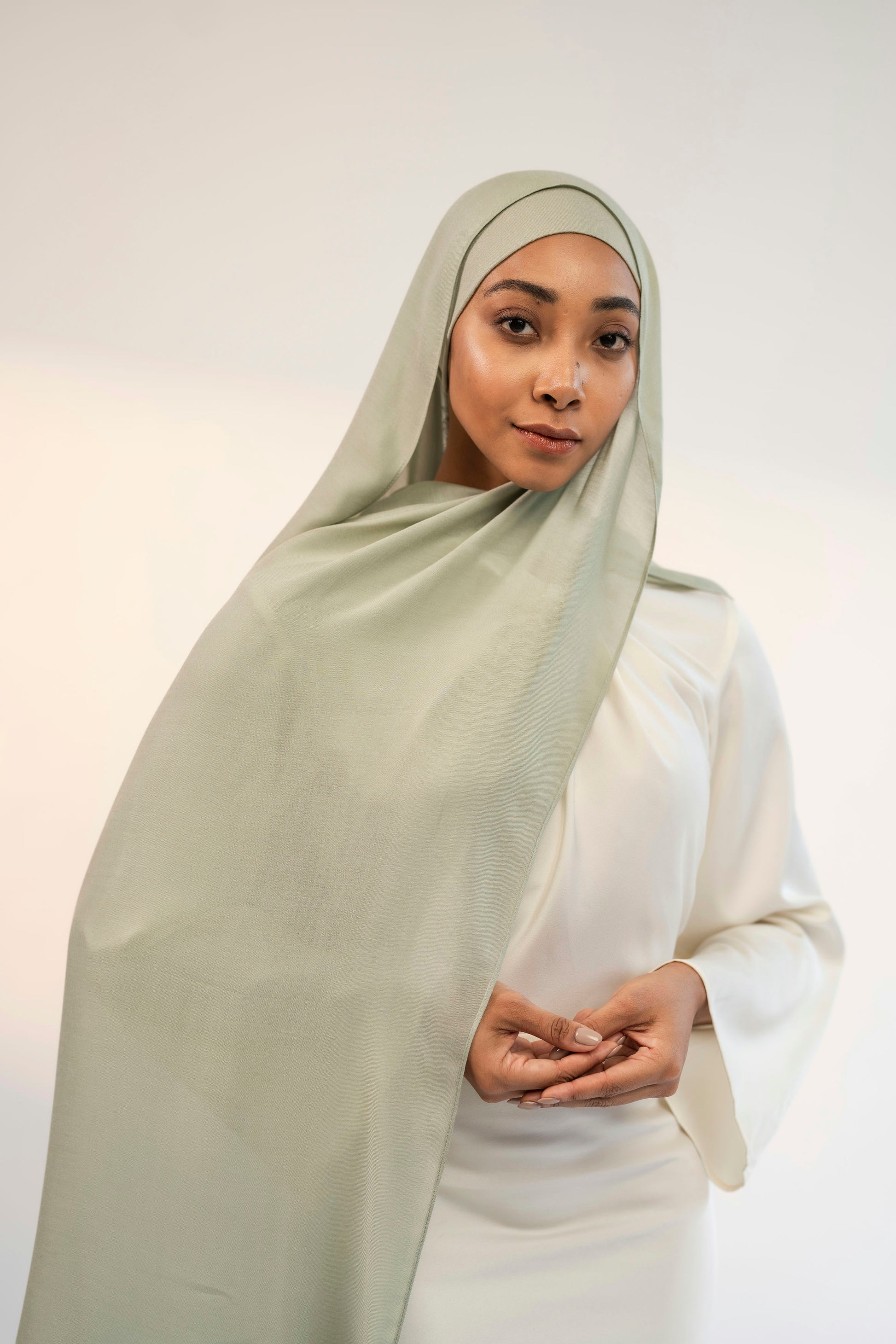 Woman wearing a light green headscarf and white outfit against a plain background 