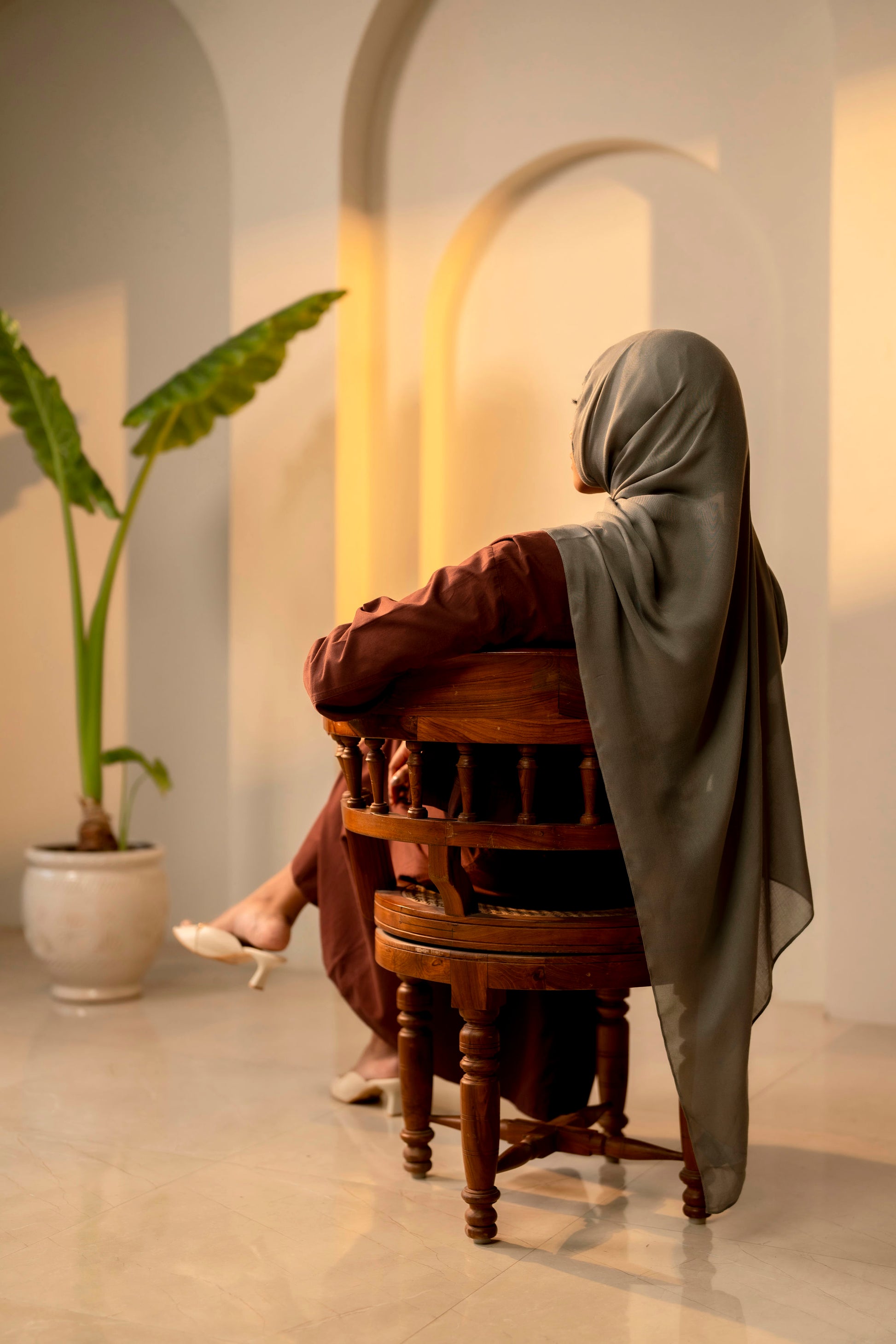 Person sitting on a wooden chair in a softly lit room with a plant in the background - sage green