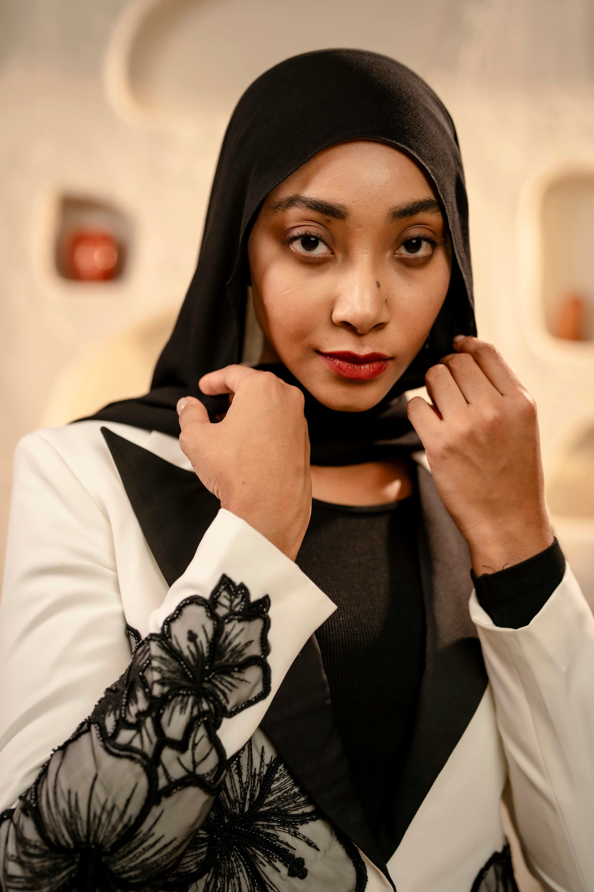Woman wearing a black hijab and white jacket with floral design indoors.