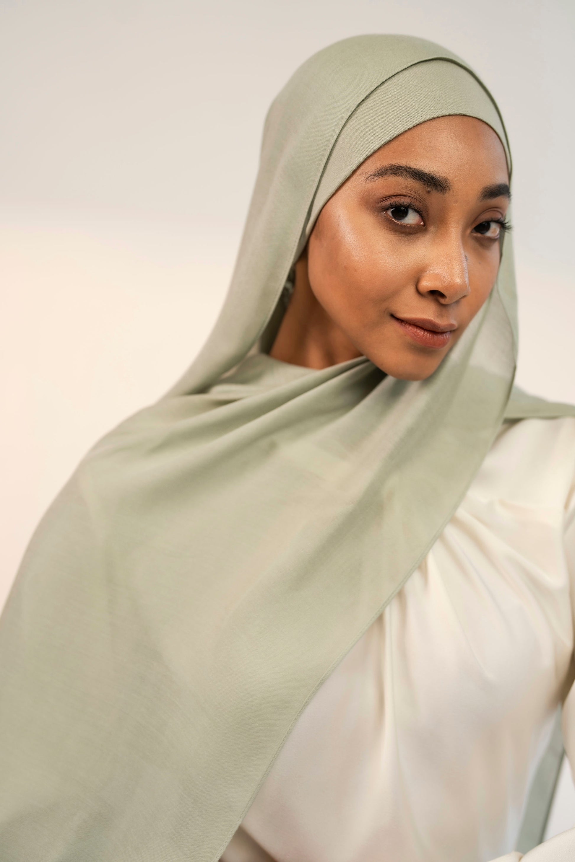 Woman wearing a matcha green hijab with a white dress against a plain background 