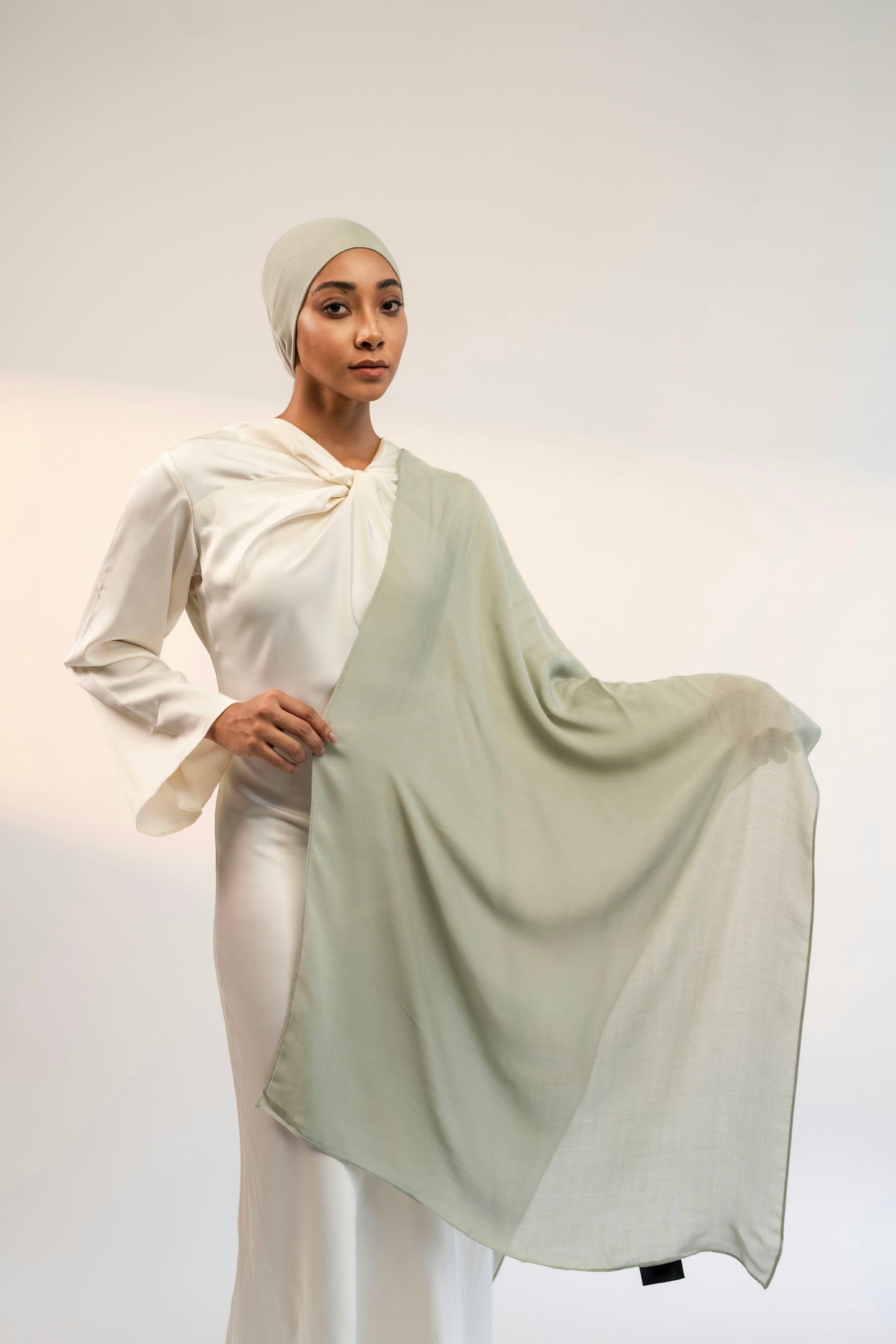 Woman wearing a white outfit with a flowing light green scarf on a plain background